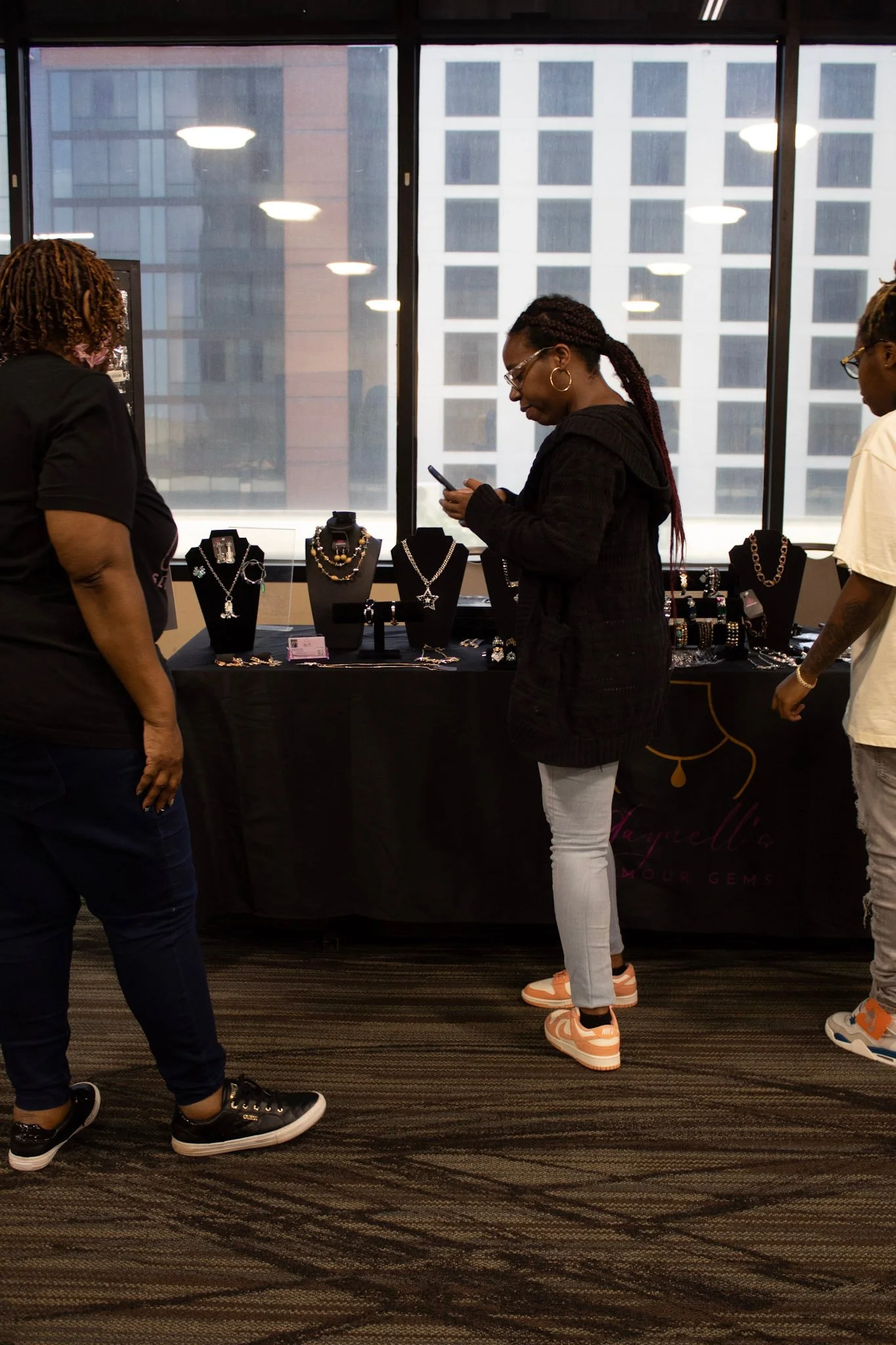 Three women browsing jewelry at an indoor market booth with large windows and city buildings in the background.