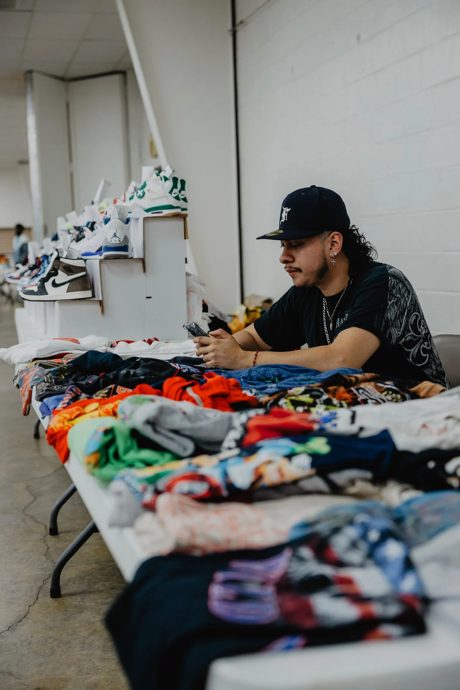 A man sitting at a table with colorful clothing and sneakers displayed in front of him, looking at his phone, at an indoor market or swap meet.