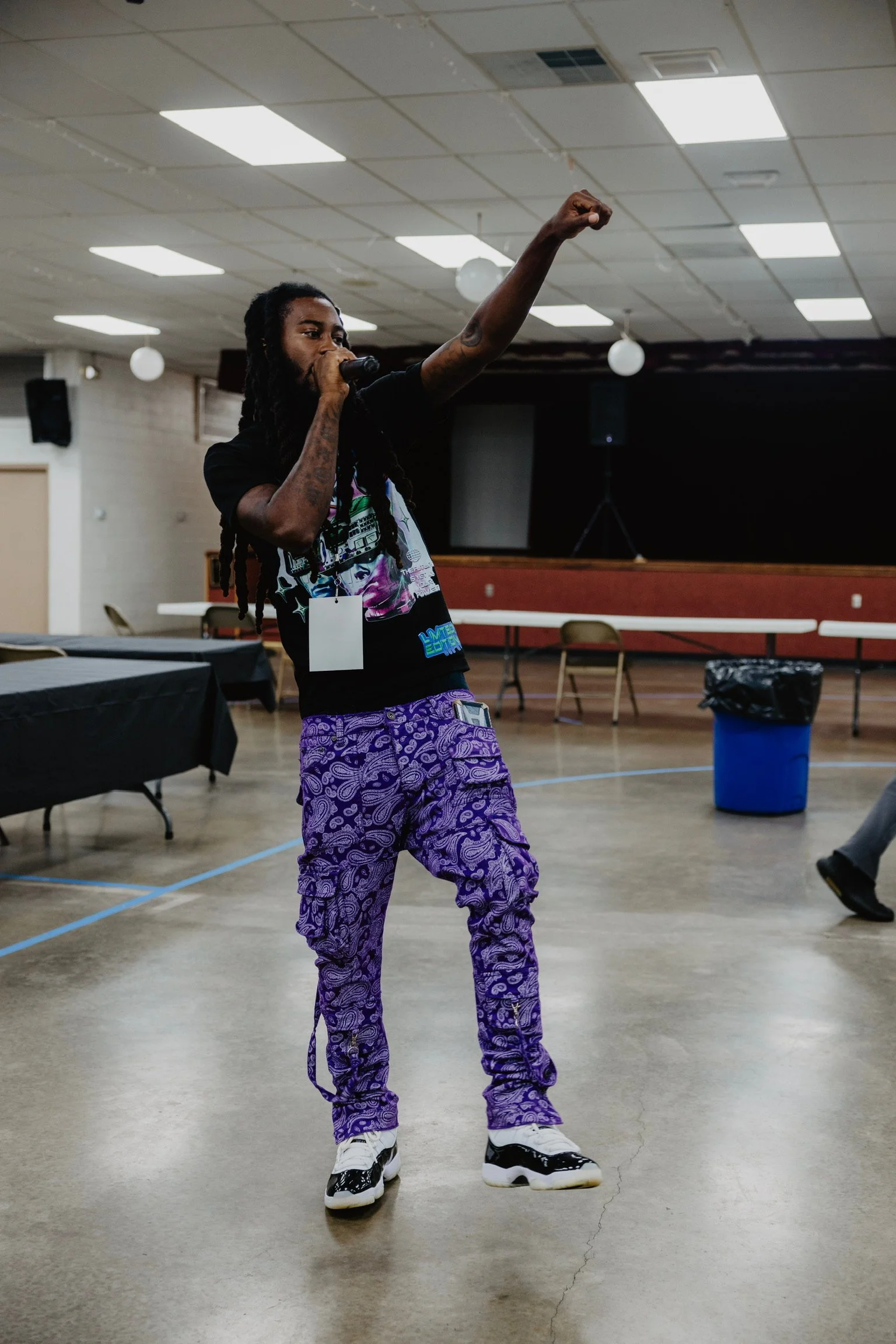 A person with long dreadlocks, wearing purple patterned pants and a black graphic T-shirt, holding a microphone in one hand and raising their fist in the air inside a large room with a stage, tables, and chairs.
