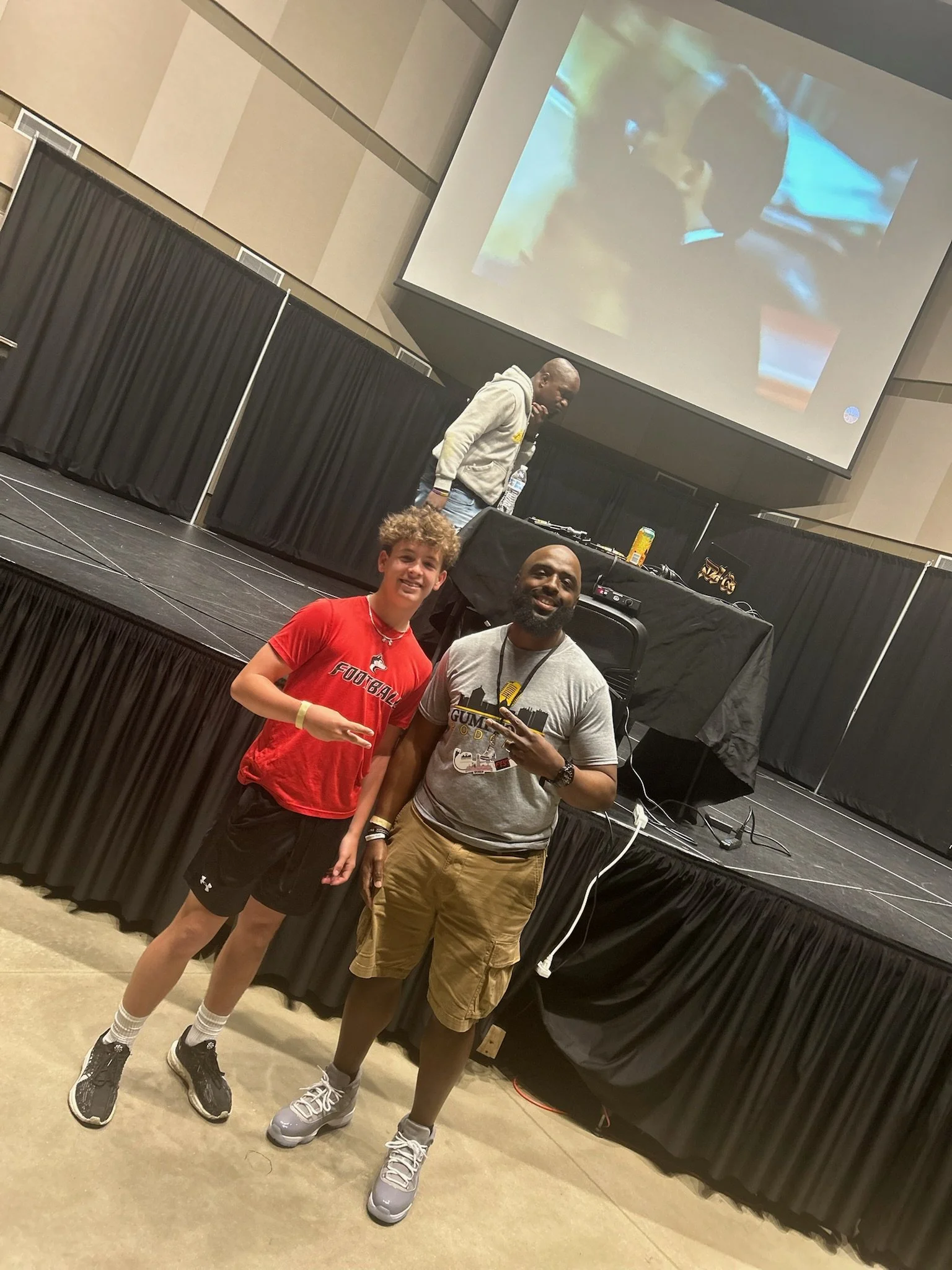 Two men and a young man posing in front of a stage with a large screen. The stage has DJ equipment and a DJ is standing behind it.