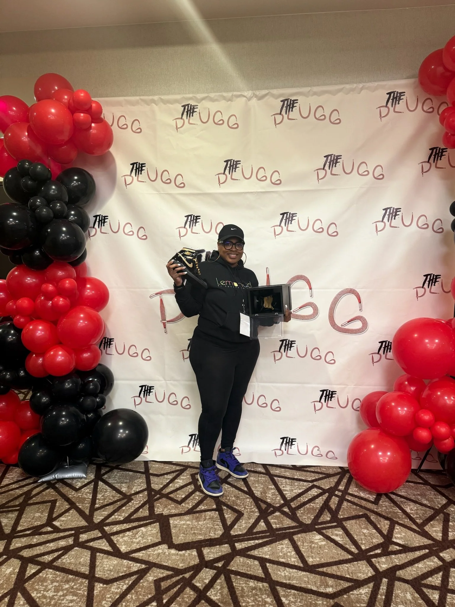 Woman standing in front of a decorated backdrop with balloons and a banner, holding a pair of shoes and a box, at an event called The Plugg.