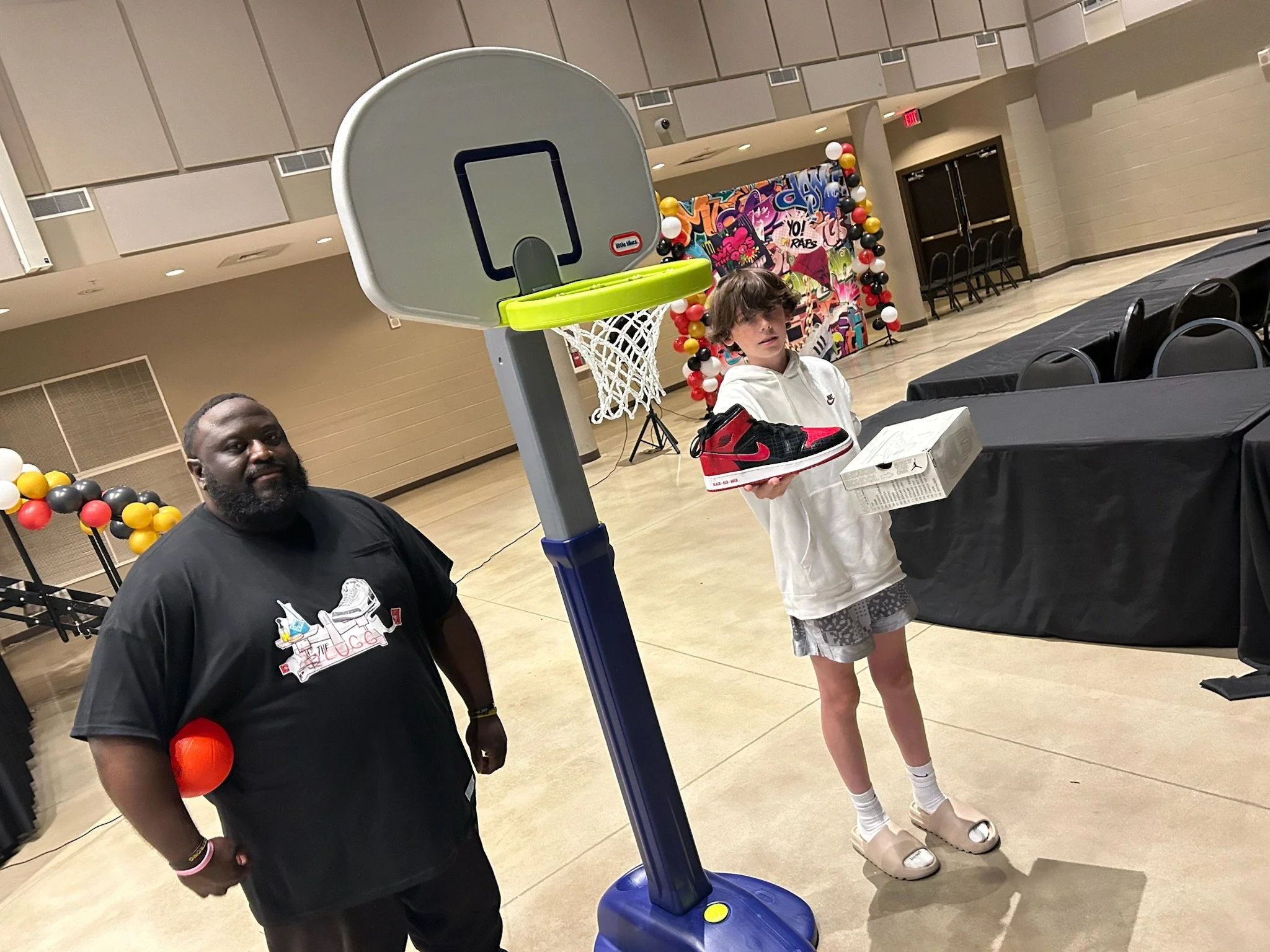 A man and a boy in a gymnasium standing near a small basketball hoop. The man is smiling and wearing a black T-shirt with a sneaker graphic. The boy is holding a sneaker and a shoebox, dressed in a white hoodie, shorts, and slide sandals. There are d