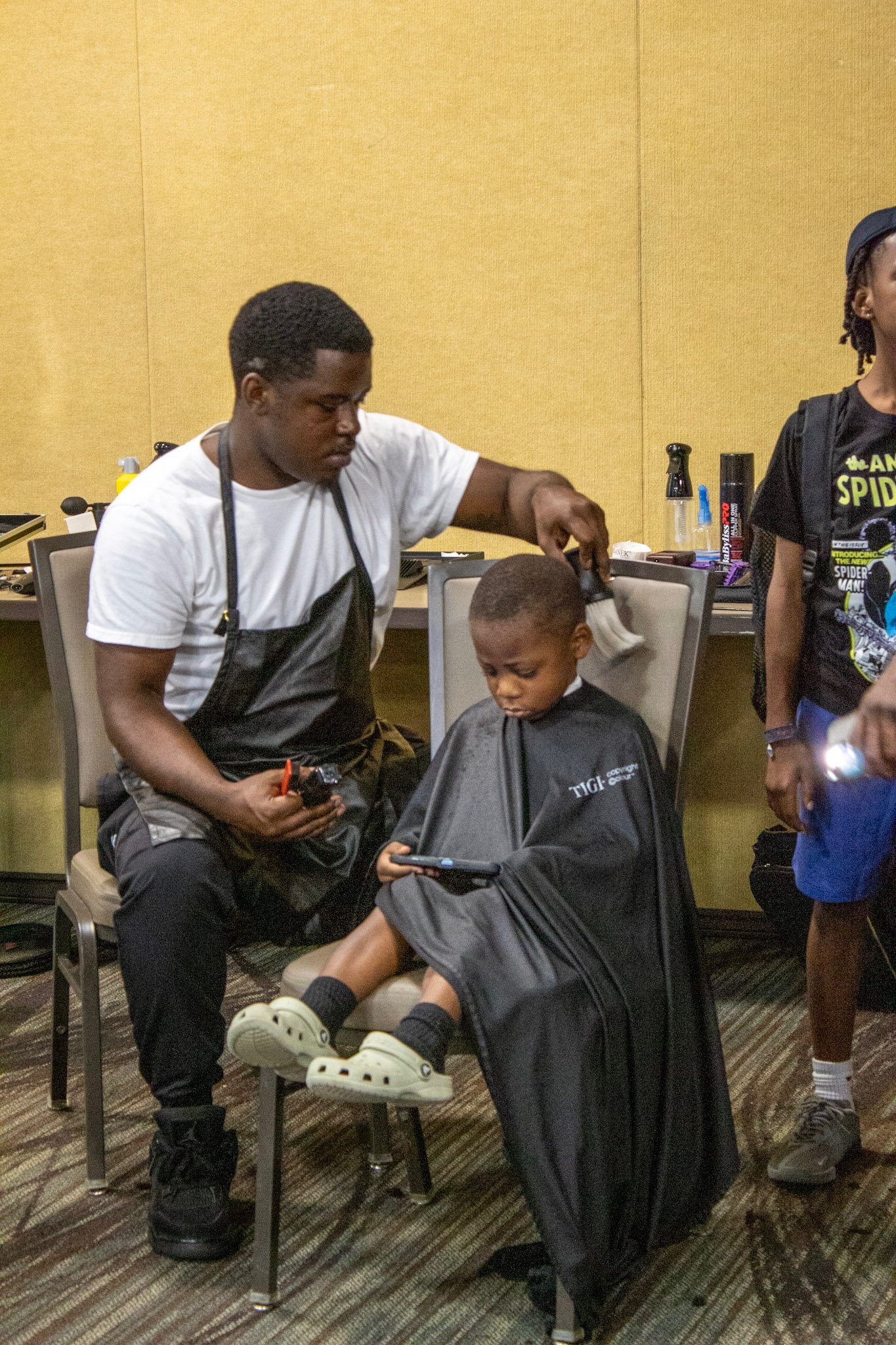 A young boy getting a haircut at a barber shop while seated on a chair, looking at a mobile device. The barber is holding a hair clipper in one hand and a brush in the other.