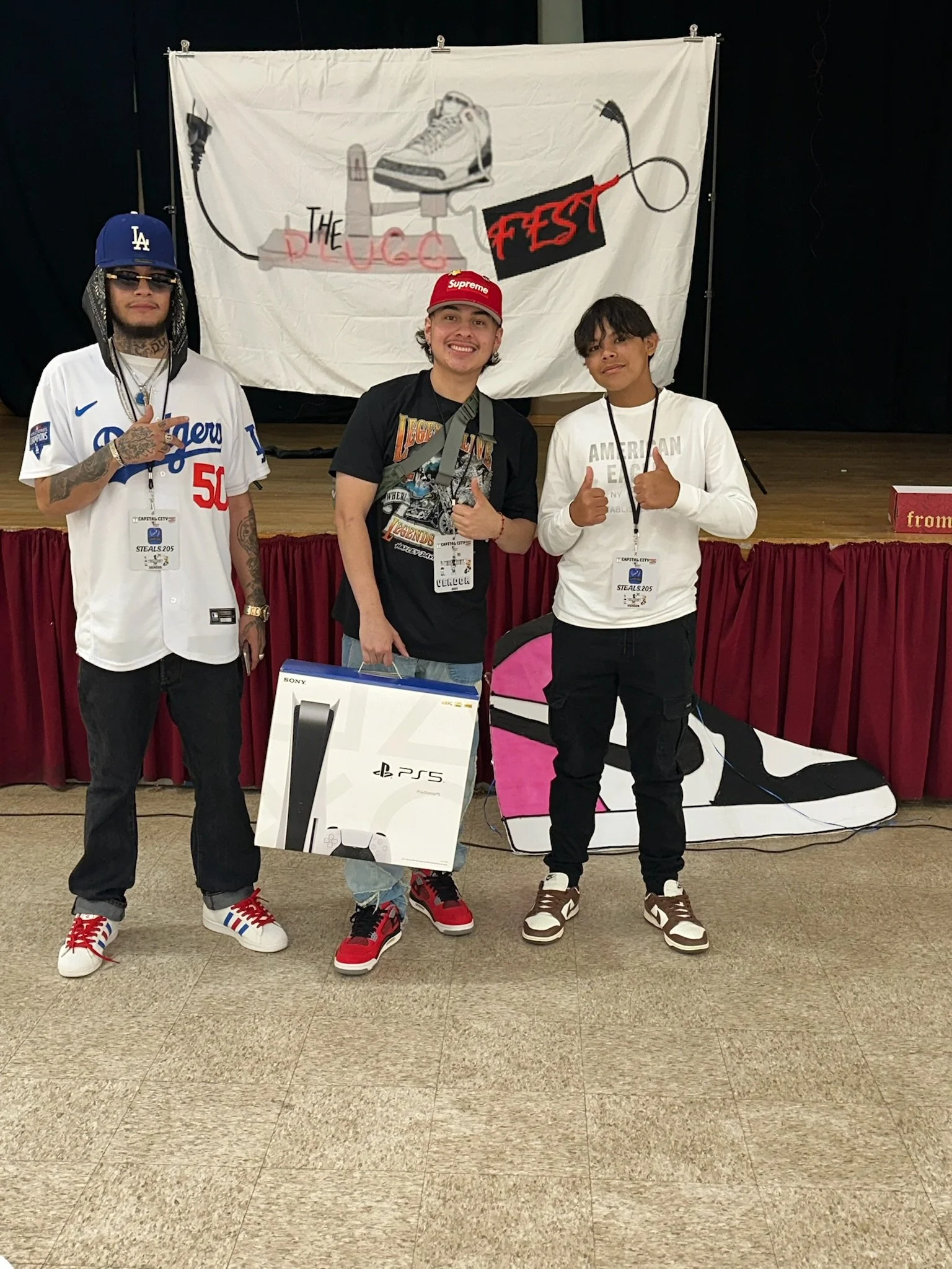 Three young men standing on a stage in front of a hand-painted banner that reads "The Lucc Fest." They are smiling and posing for the photo, with the middle person holding a PlayStation 5 box. The man on the left is making a peace sign, wearing a Los