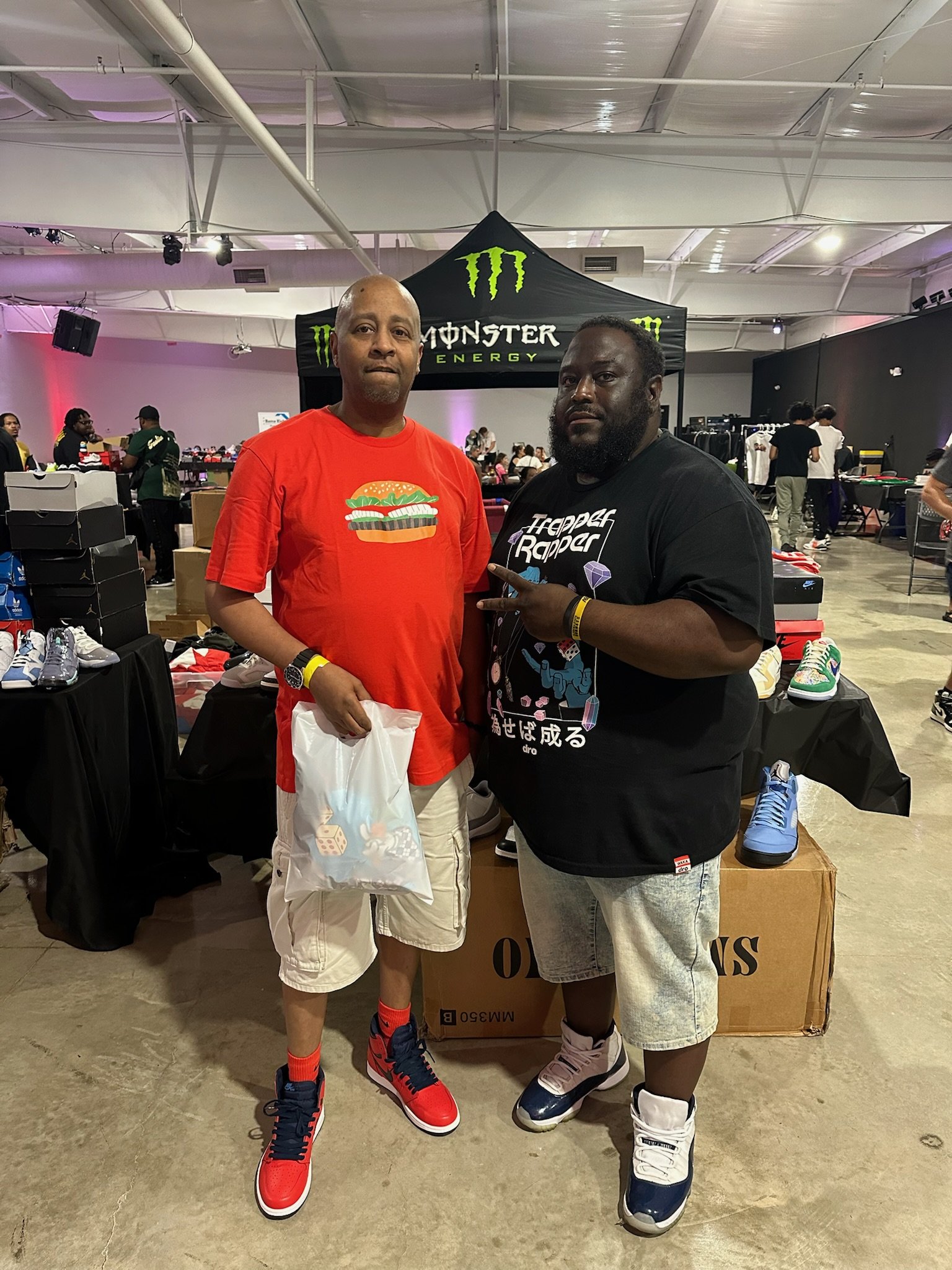 Two men standing inside a sneaker or sneaker-related event, with tables of shoes and a black Monster Energy stand in the background. The man on the left is wearing a bright red t-shirt with a burger graphic, beige shorts, and red and blue sneakers. H