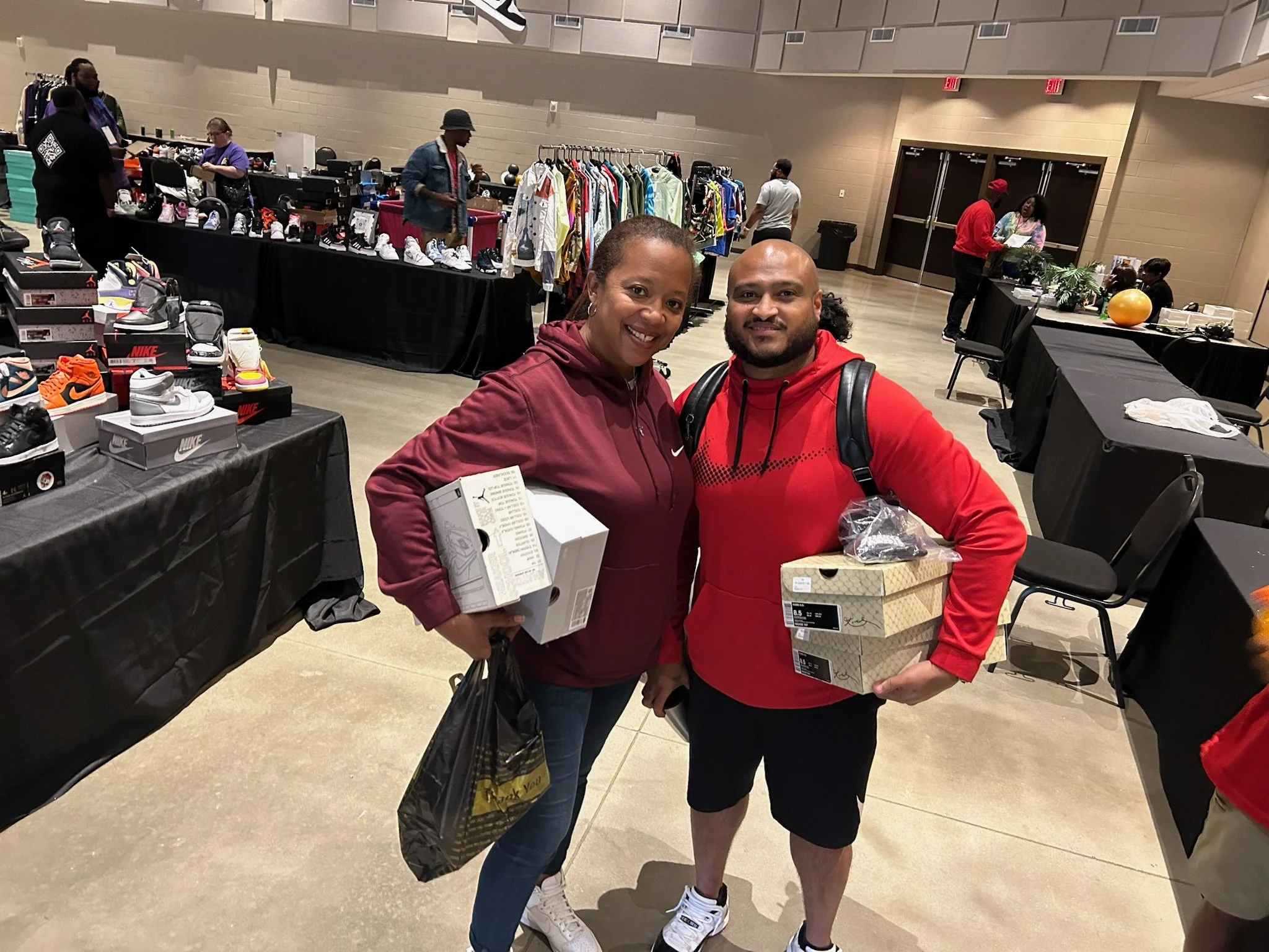 A smiling woman and man standing together at a shopping event, holding shoe boxes. The woman is wearing a maroon hoodie and jeans, and the man is wearing a red hoodie and black shorts. Background shows tables with shoes, clothing racks, and other sho