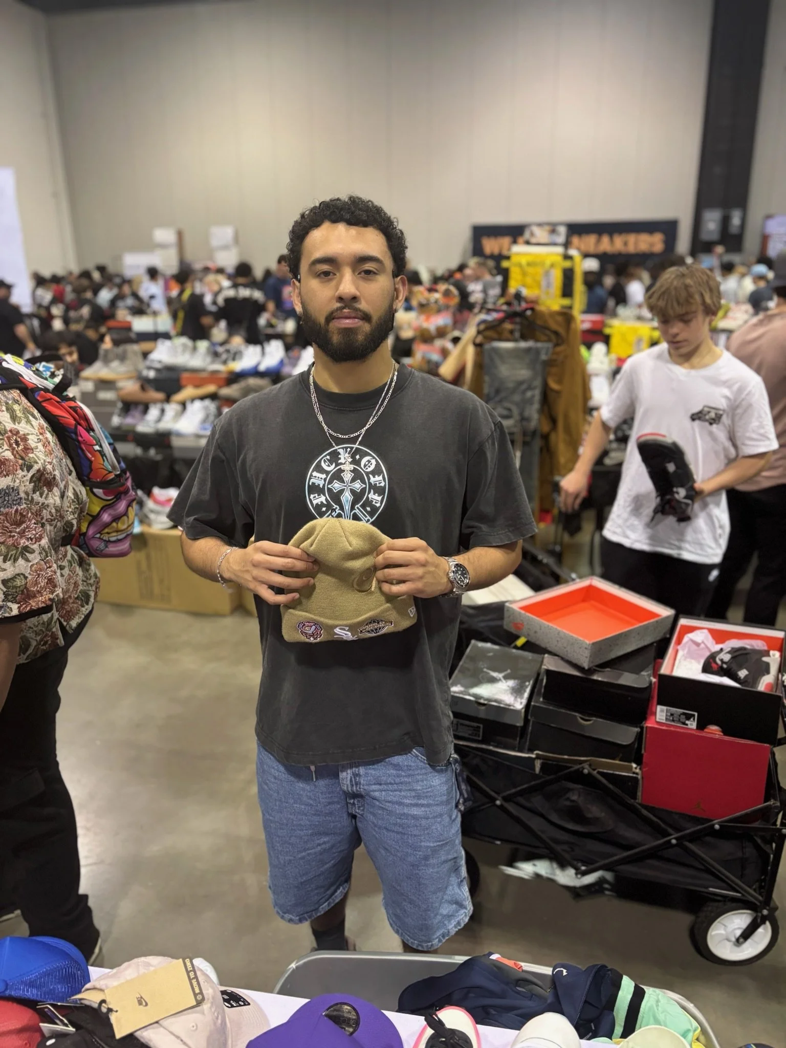 A young man with a beard and curly hair, wearing a black t-shirt and blue shorts, stands in a crowded indoor marketplace or thrift store. He holds a beige beanie hat decorated with patches. There are boxes of sneakers and other items on tables and ca