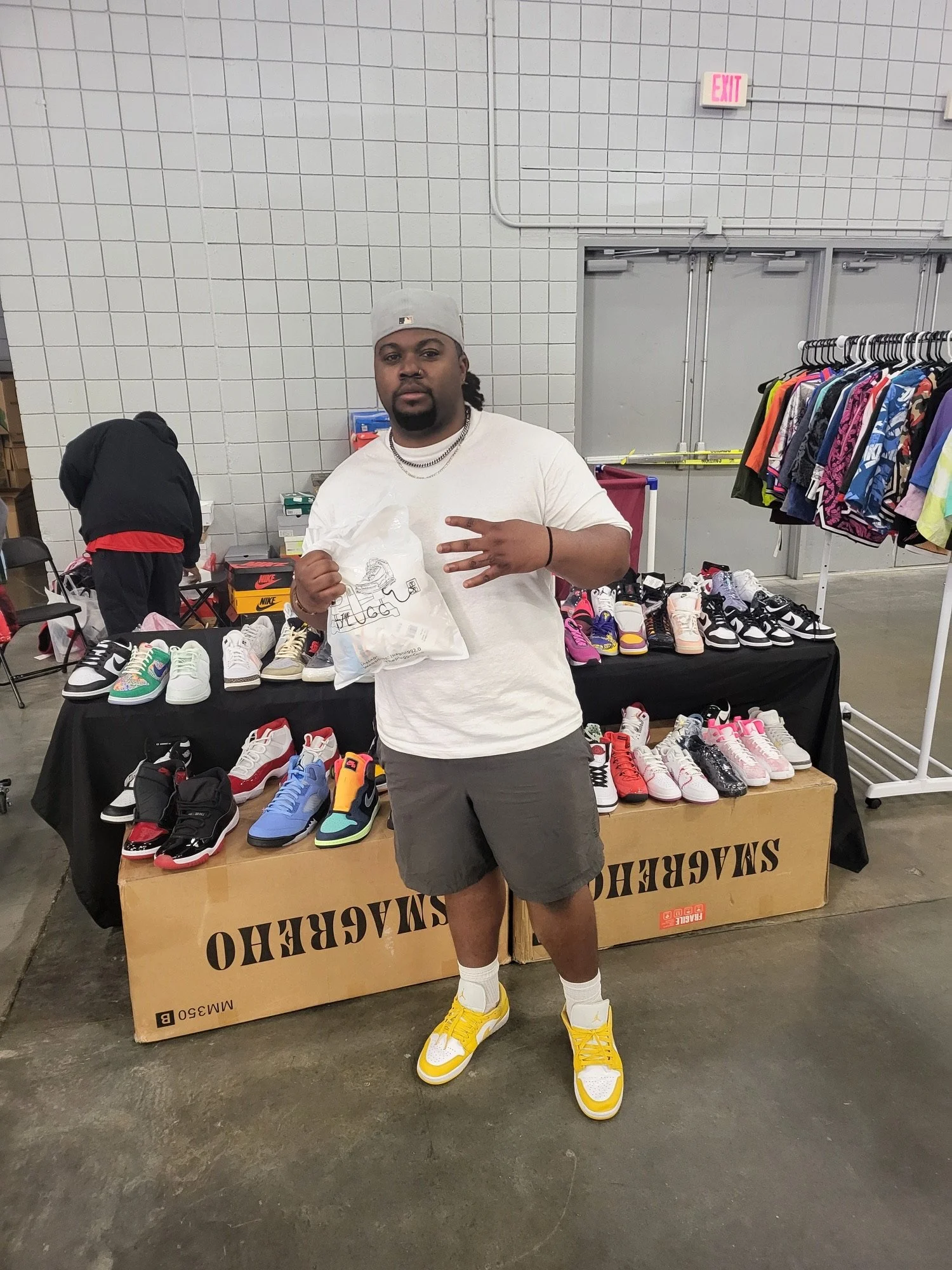 A man posing in front of a table of sneakers at an indoor sneaker sale event, holding a plastic bag and making a peace sign with his right hand, wearing a white shirt, gray shorts, yellow and white sneakers, and a gray cap.