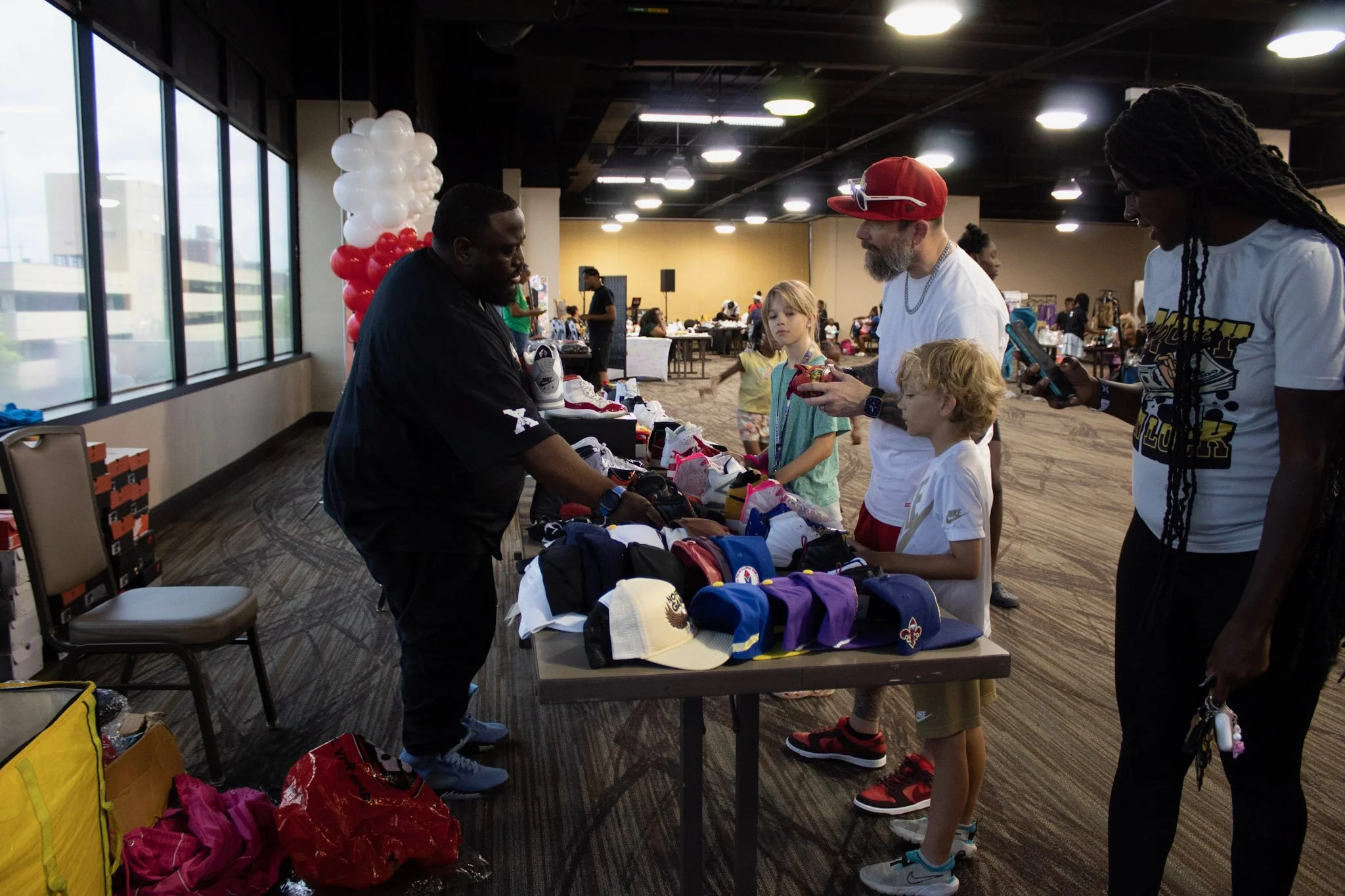 People shopping for sneakers at an indoor event with tables displaying shoes, balloons, and other vendors in the background.