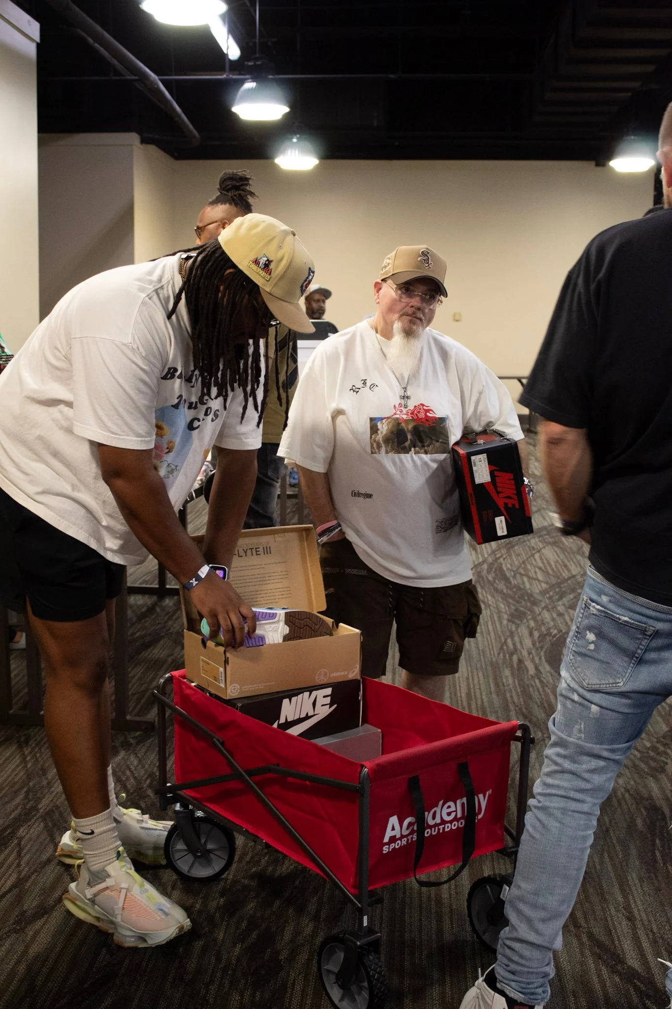 People shopping for sneakers at an indoor store, with a red cart labeled 'Academy Sports + Outdoors' filled with shoeboxes, including Nike boxes, and one person holds a Nike shoe box.