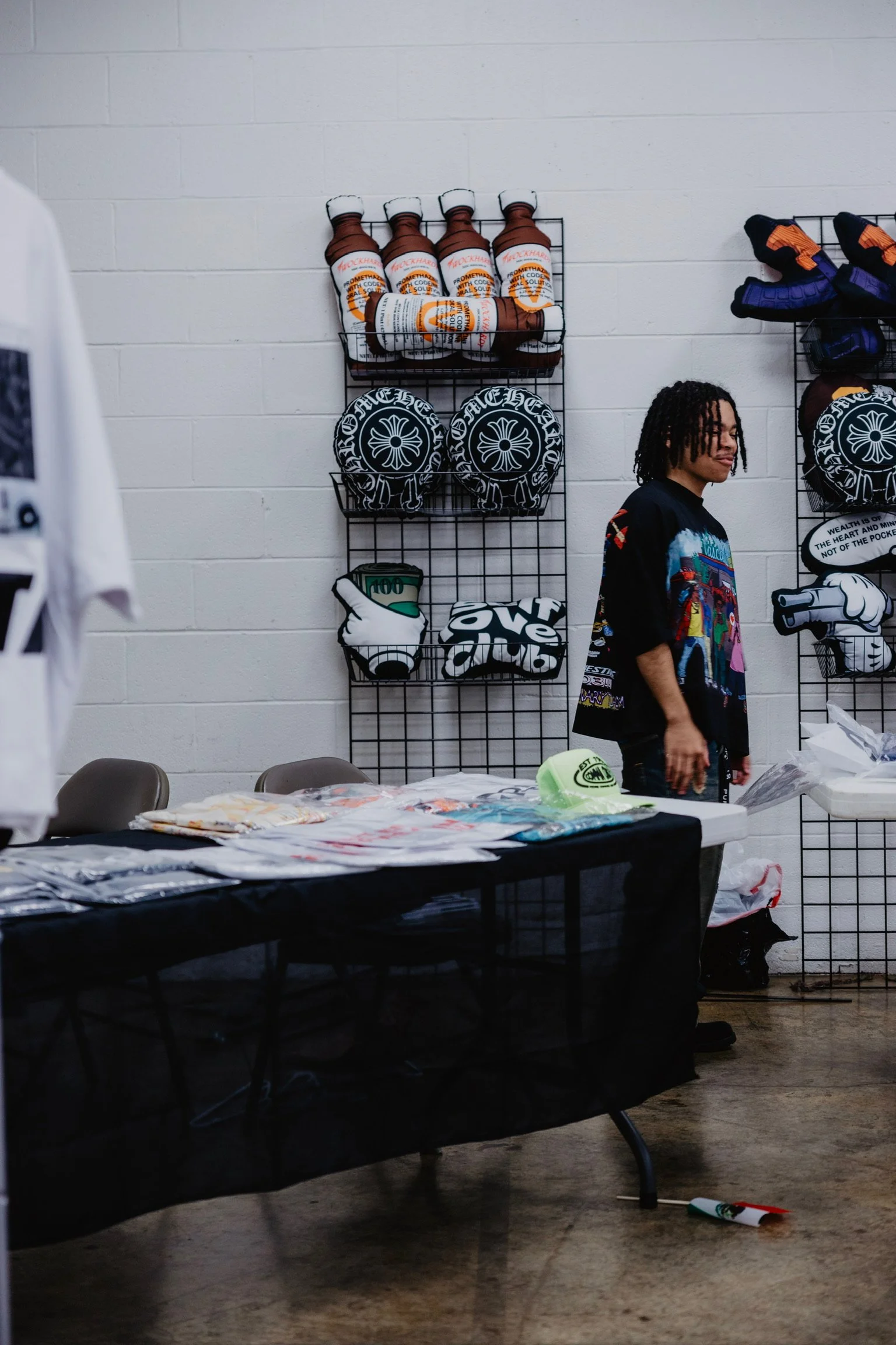 A man with dreadlocks standing behind a black table with merchandise, in a room with a white cinder block wall, displaying various fashion items and wall-mounted accessories.