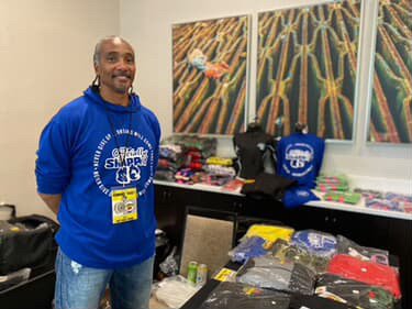 Man standing in front of a table with various items, wearing a blue hoodie, at an indoor event with artwork on the wall behind.