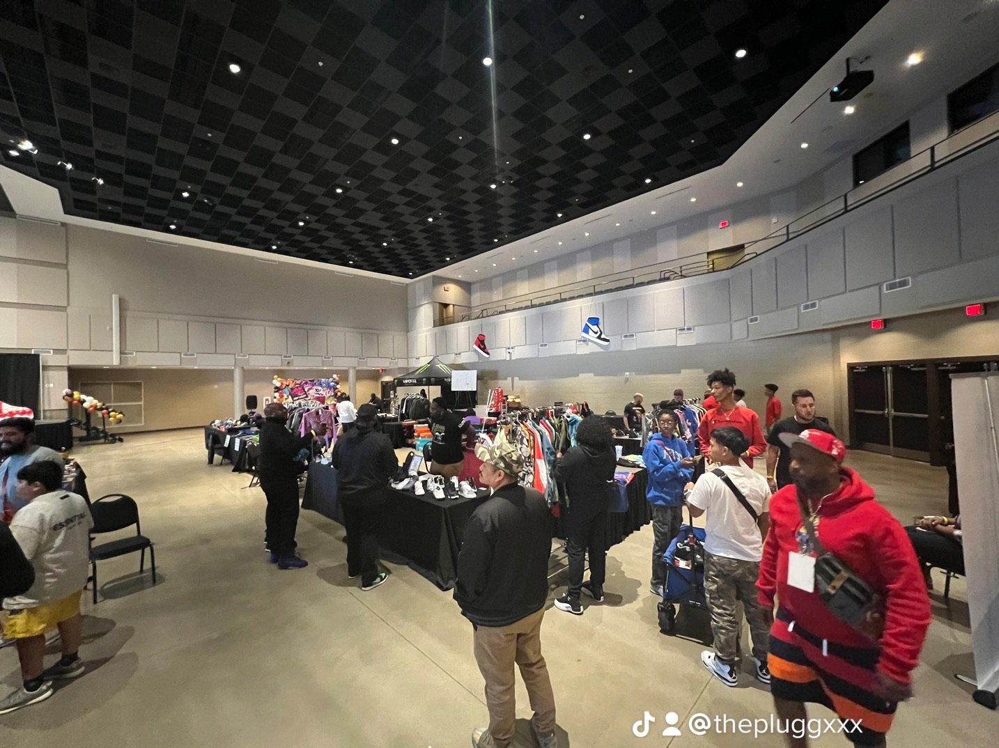 An indoor market or event with people browsing clothing and accessories at various tables, with some wearing casual streetwear or sportswear, in a spacious hall with high ceilings and overhead lighting.