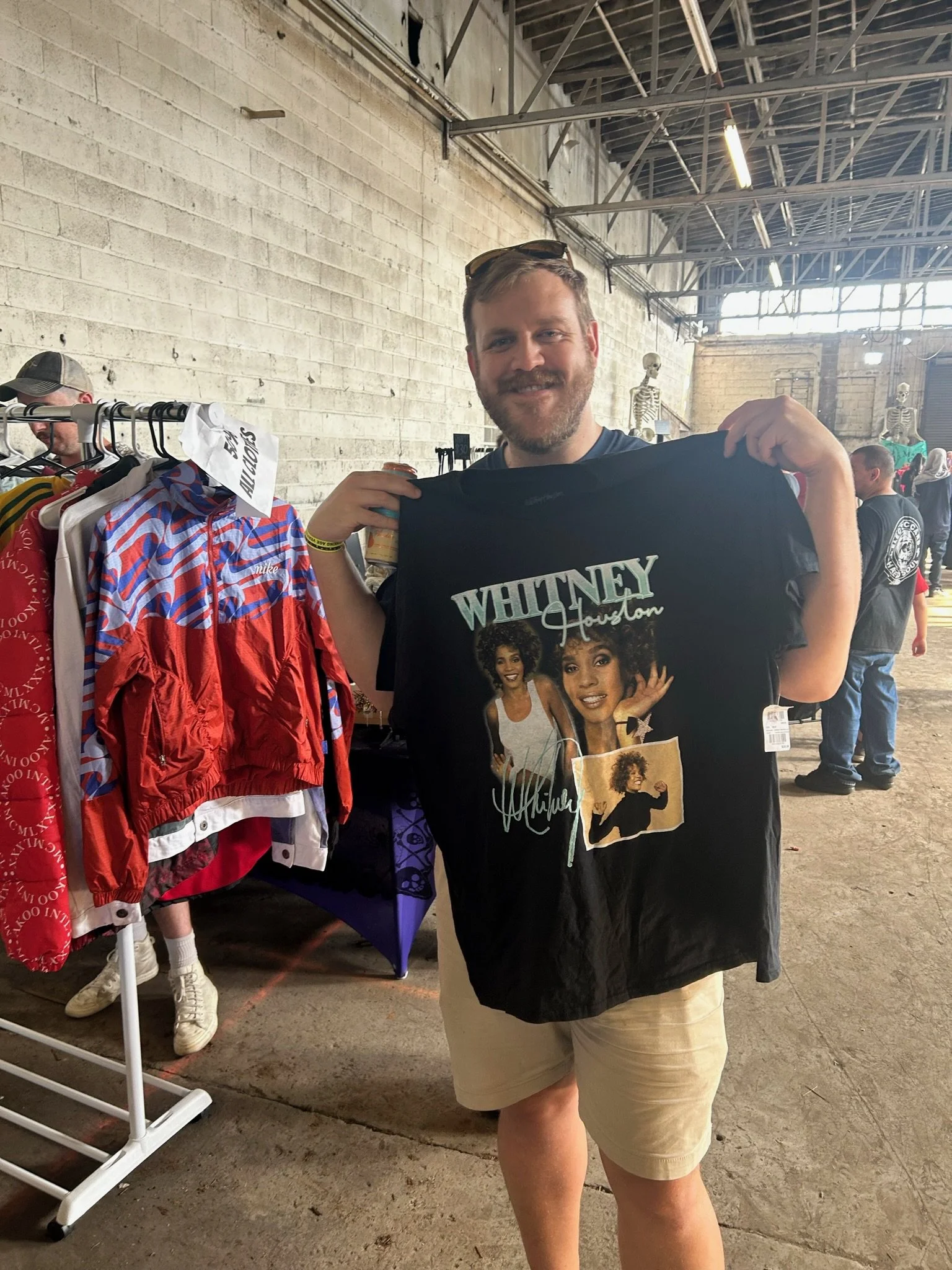 A man holding a Whitney Houston T-shirt at a flea market or indoor sale.