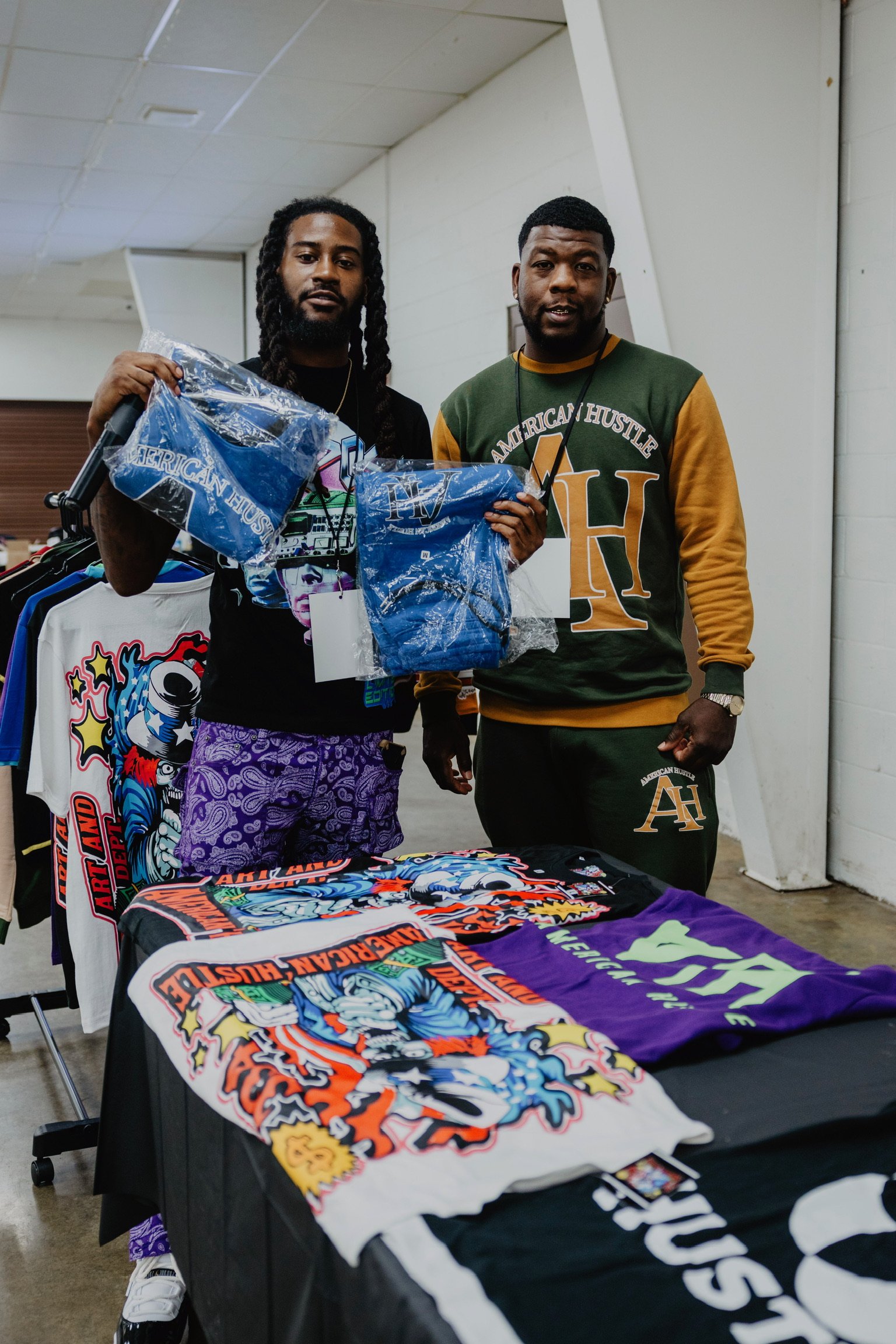 Two men standing behind a table filled with colorful graphic T-shirts, holding packaged clothing items, in an indoor setting.