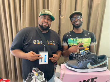 Two men smiling, standing behind a table with Nike shoes, one holding a badge, the other pointing at the shoes.