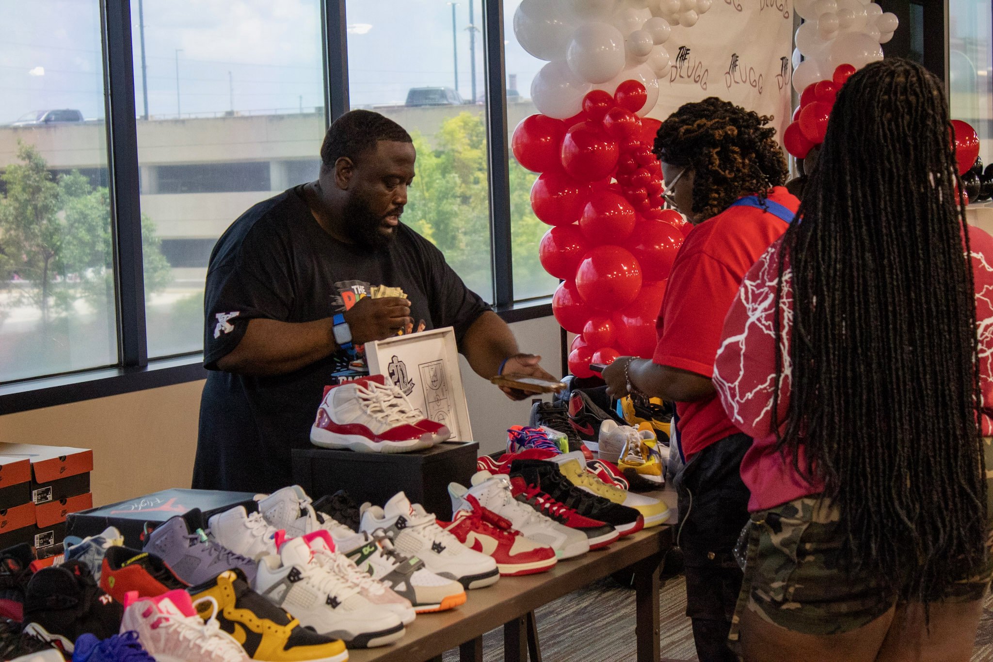A man selling sneakers at a booth decorated with red, white, and black balloons, with two women browsing shoes displayed on the table.