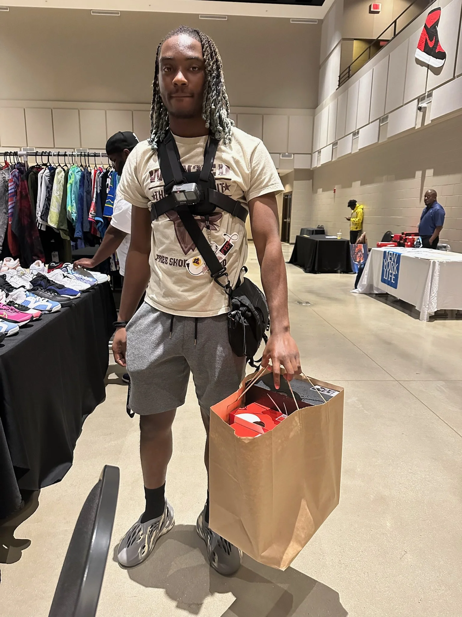 A young man with dreadlocks wearing a beige T-shirt, gray shorts, and sneakers holding a brown shopping bag with Nike shoes inside at a sneaker event or sale.