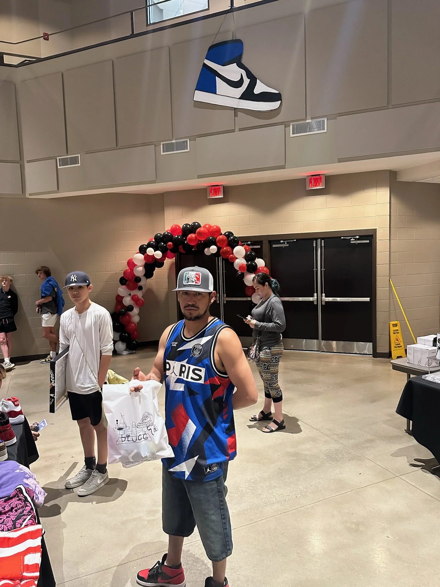 A man wearing a Paris basketball jersey and a cap holding a bag, standing in a decorated event space with balloon arch and sneaker signage.