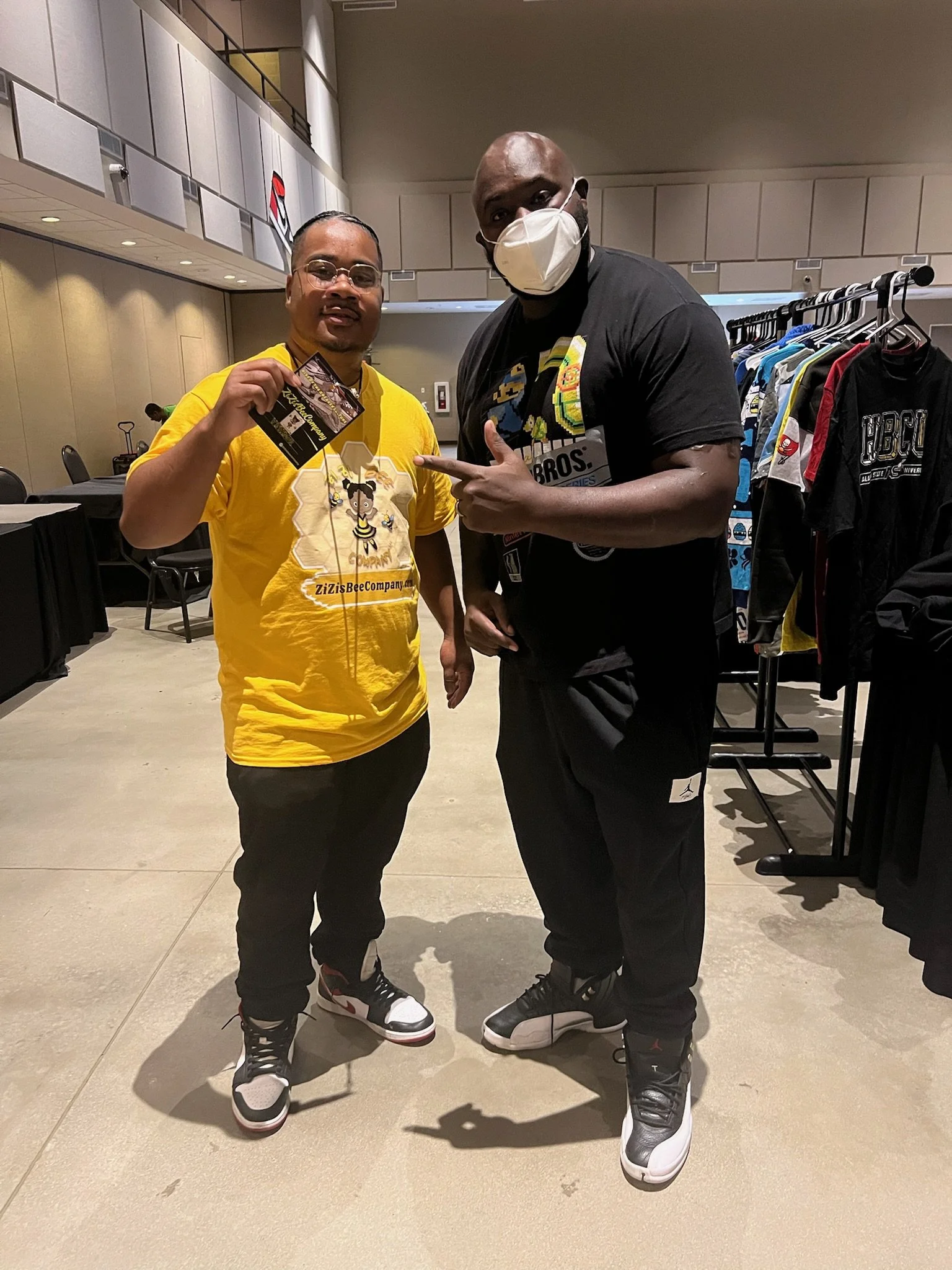 Two men standing indoors in front of a clothing rack. One man is holding a ticket or card and wearing a yellow T-shirt, the other is wearing a black T-shirt with a mask and pointing at the man in yellow.