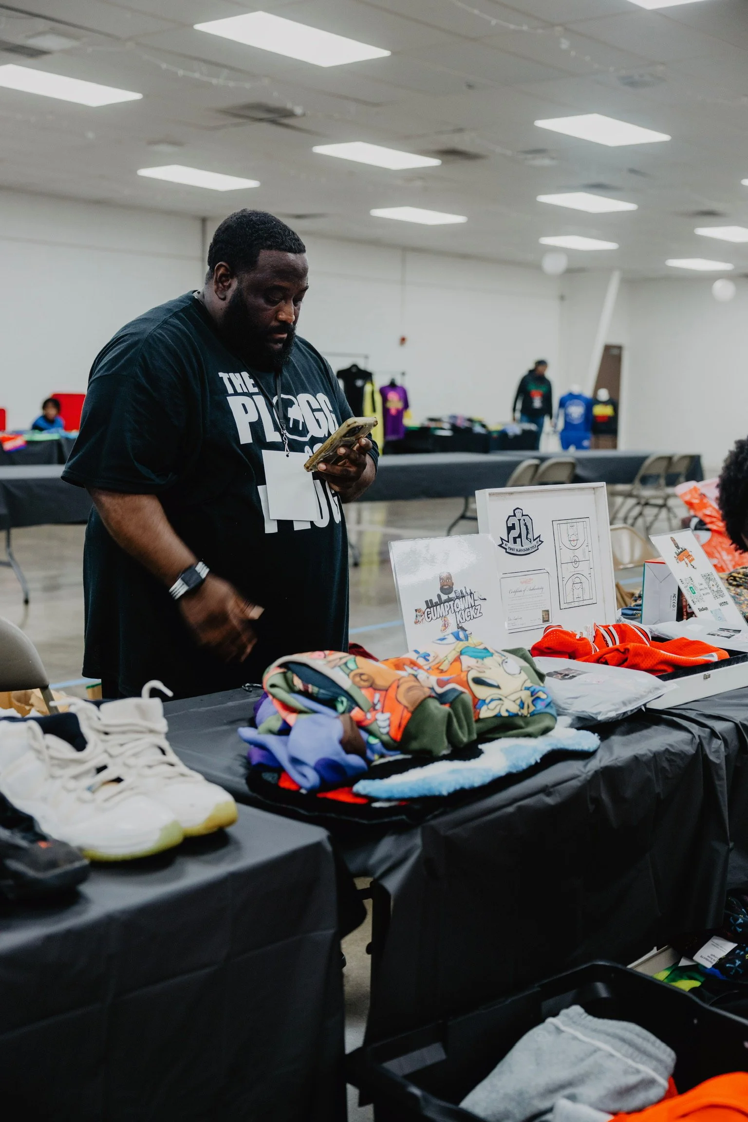 A man standing at a table with various items like clothes, shoes, and framed pictures inside a large indoor space, possibly at a marketplace or event.