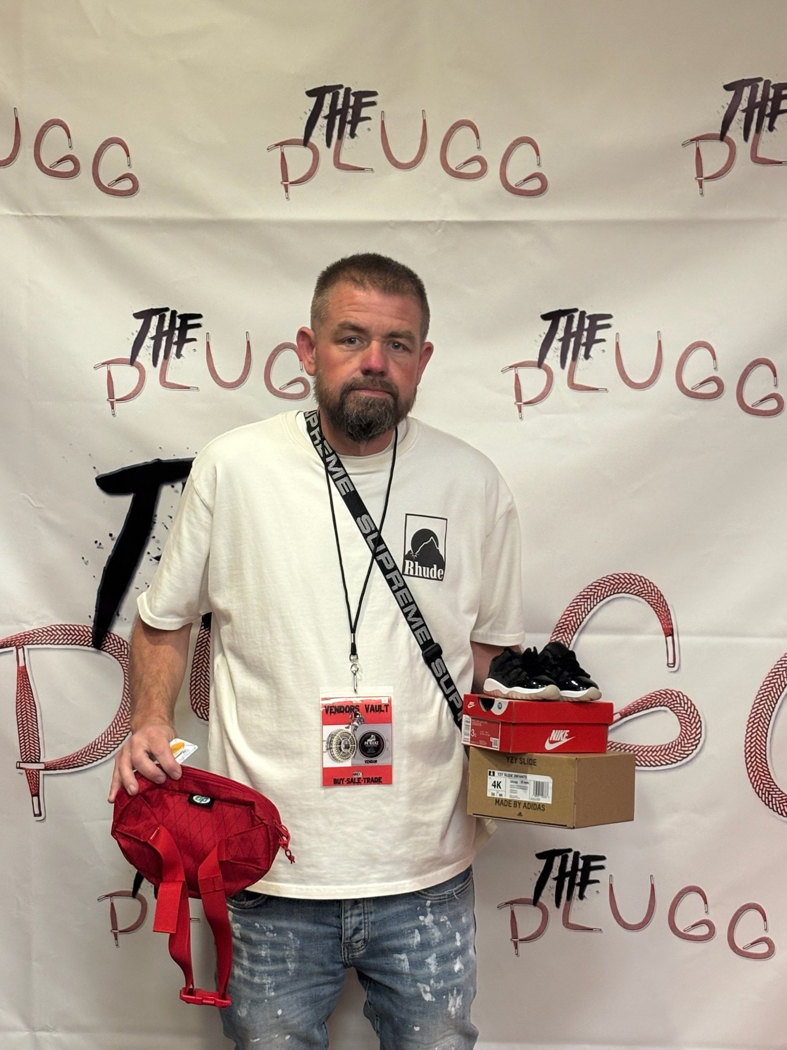 A man with a beard and mustache holding shoe boxes, standing in front of a backdrop with the words 'The Plugg' repeated on it. He has a red bag in his left hand and wears a white T-shirt with a Rhude logo.