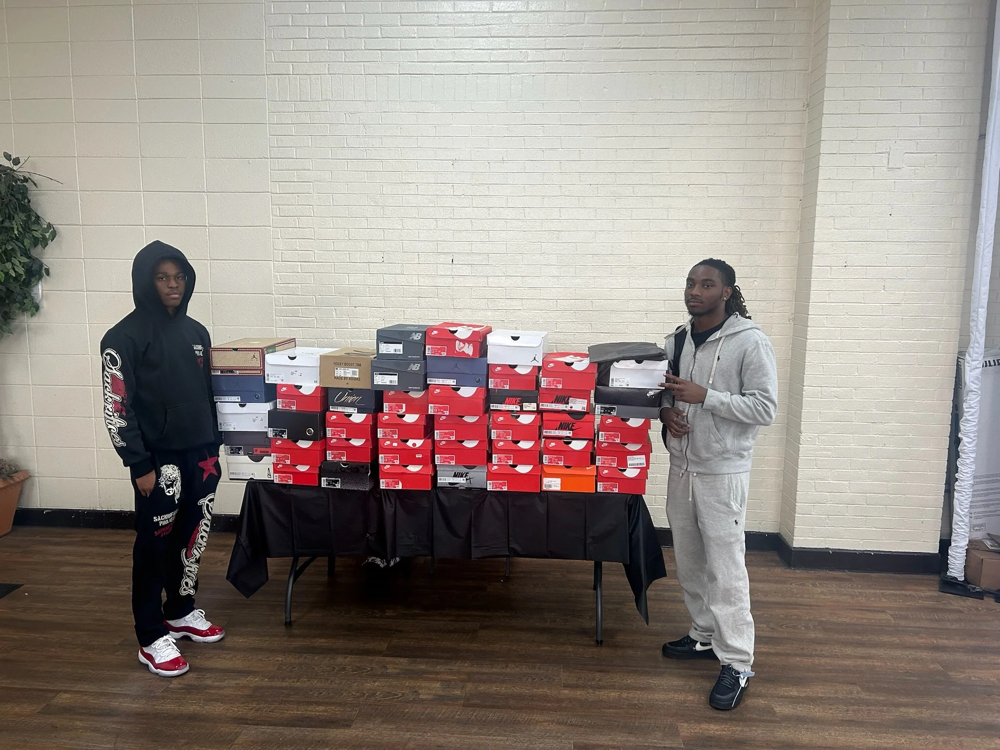 Two young men standing next to a table with multiple sneaker boxes, against a white brick wall. One man on the left is wearing a black hoodie and black pants, and the man on the right is dressed in a gray hoodie and gray sweatpants.