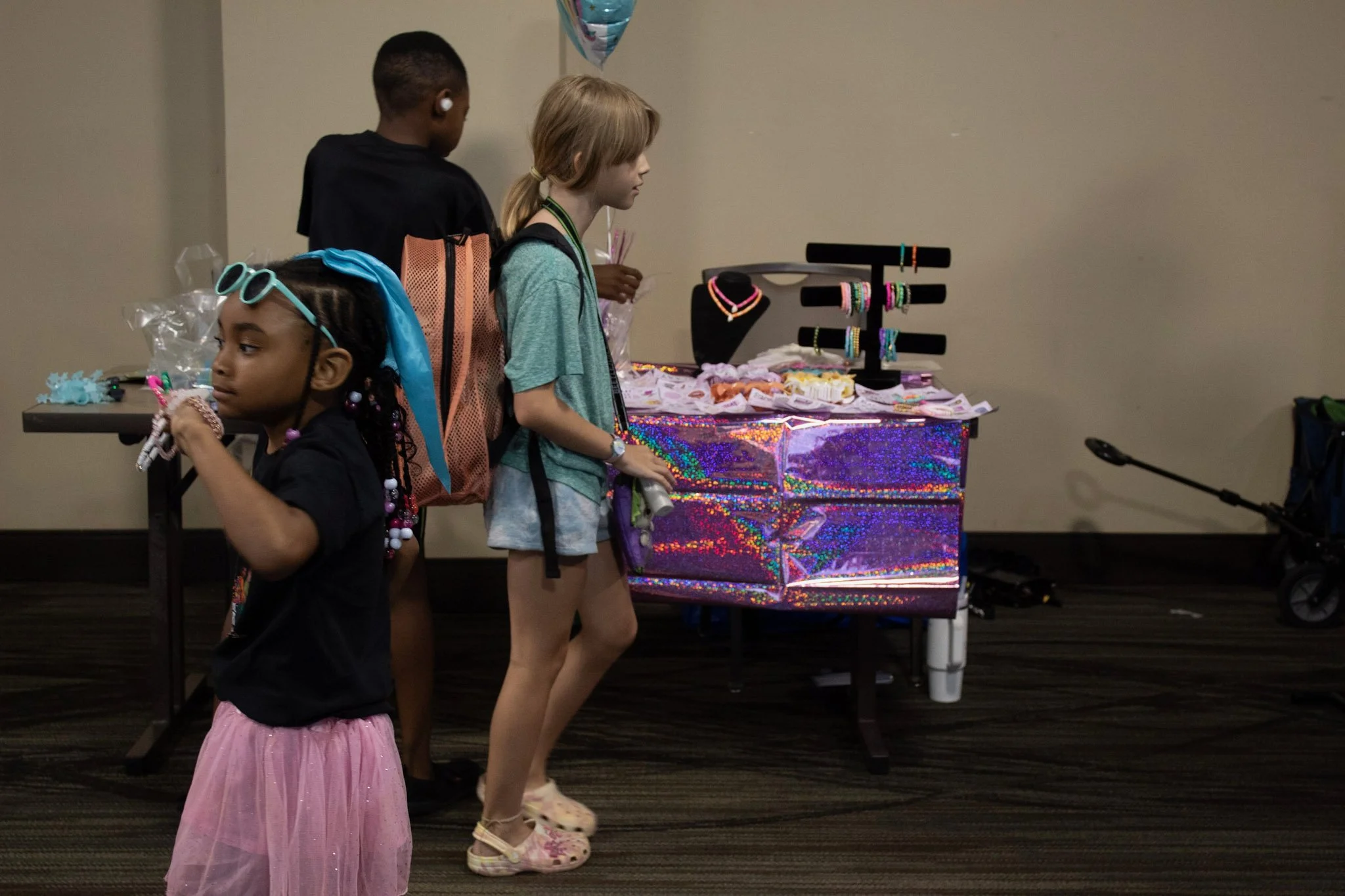 Three children standing in front of a display table with jewelry and accessories, in an indoor setting.