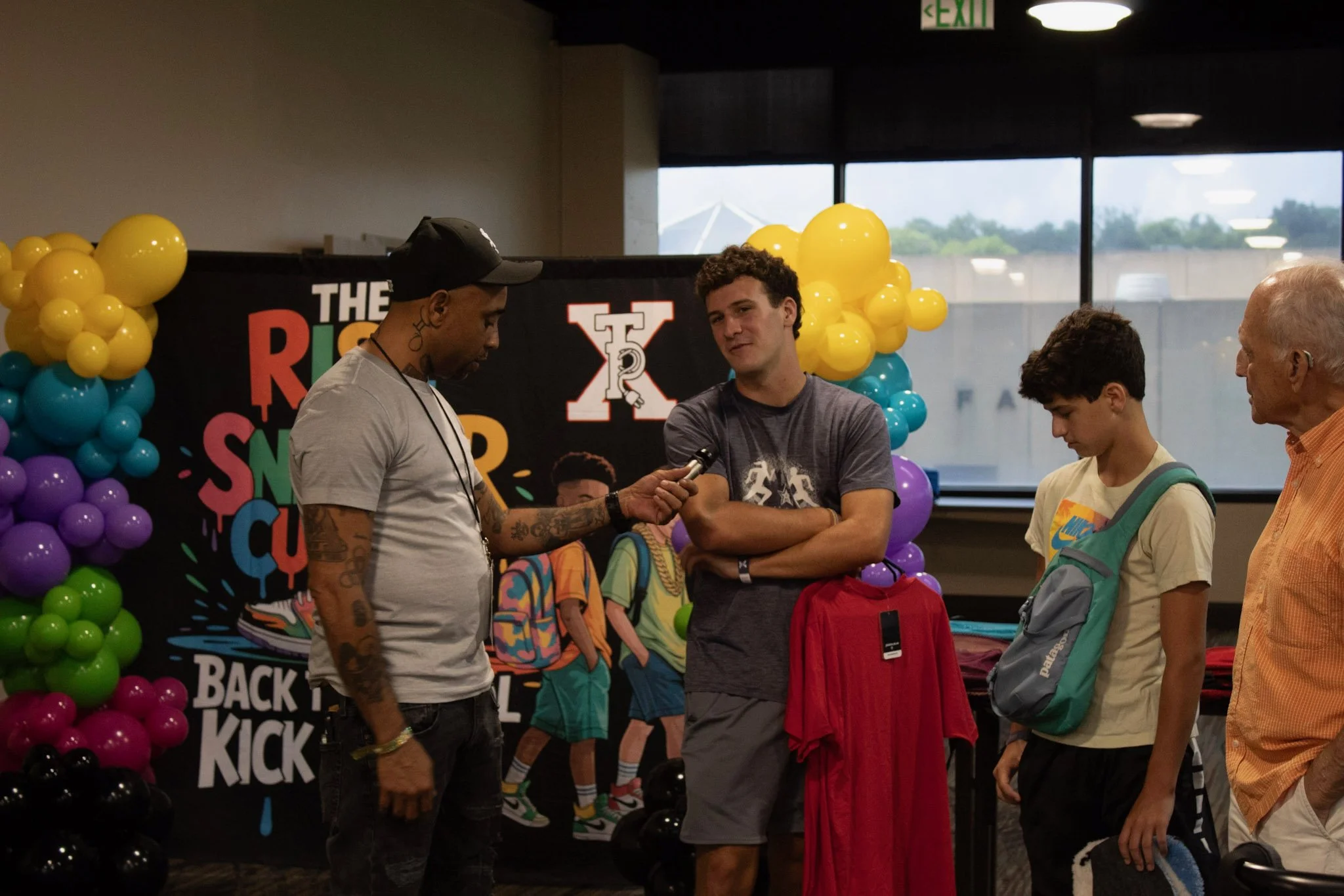 A group of people being interviewed at an event with colorful balloons and a backdrop that reads 'The Rise Up SnS Back to Kick'.