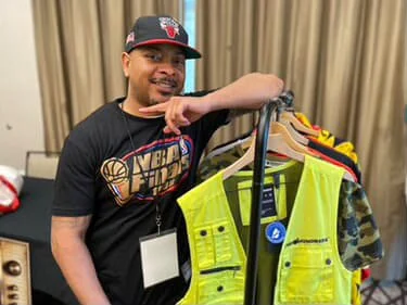 Man with a black cap and T-shirt smiling, holding a rack of high-visibility safety vests and clothing.