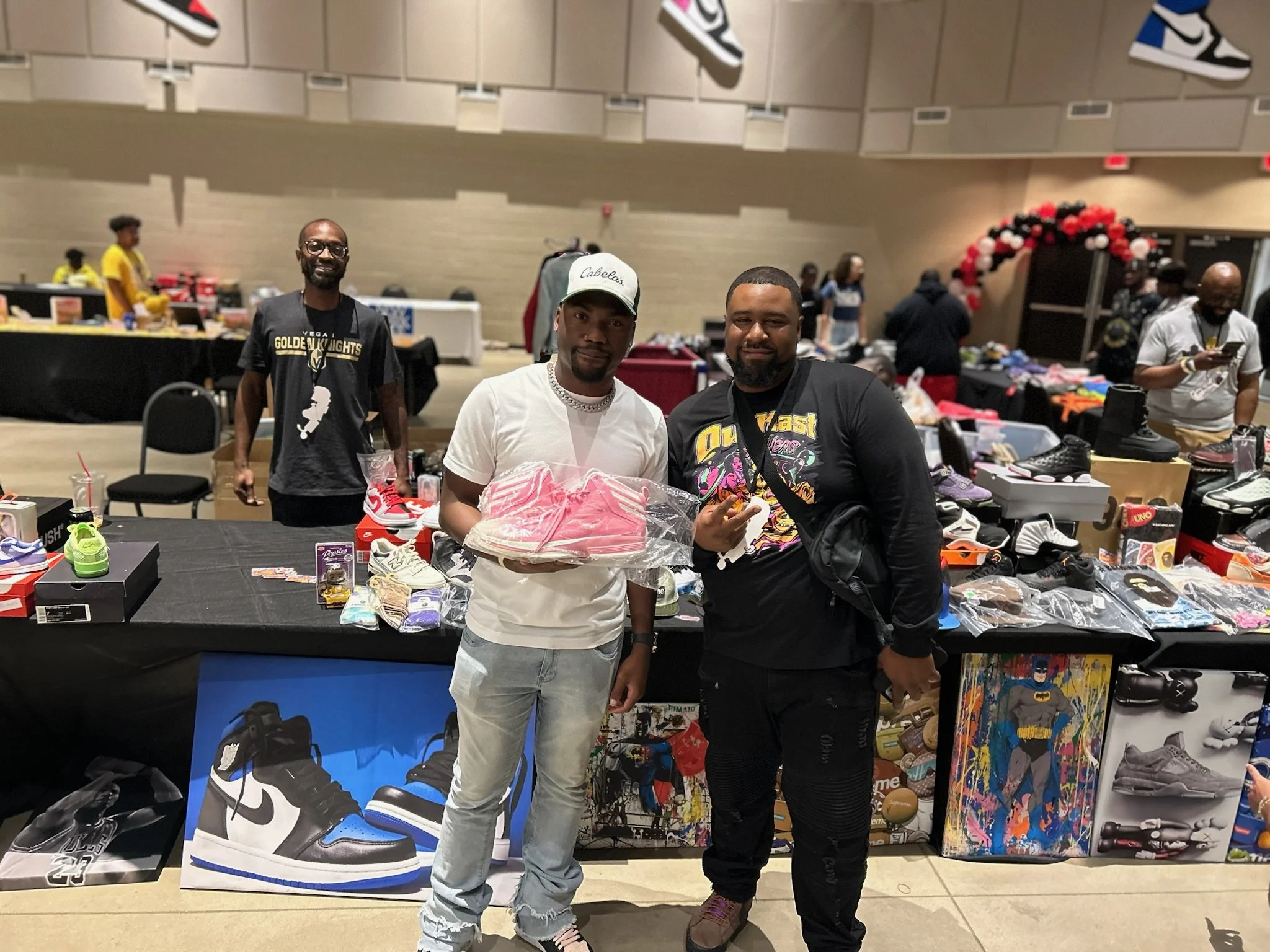 Three men at a sneaker and collectible toy booth at a convention or marketplace, with tables displaying sneakers, toys, and collectibles, and people browsing in the background.