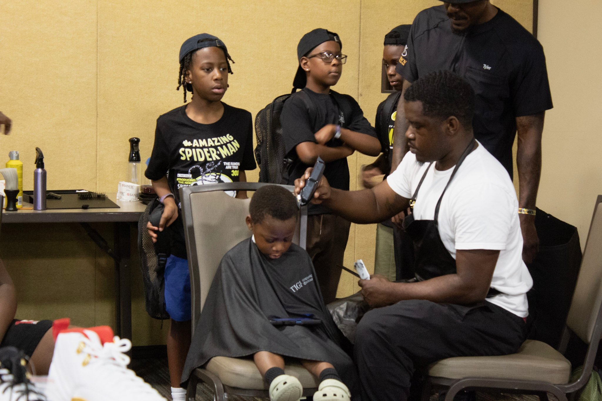 A barber trims a young boy's hair while five children stand nearby, observing the haircut in a salon or barber shop.