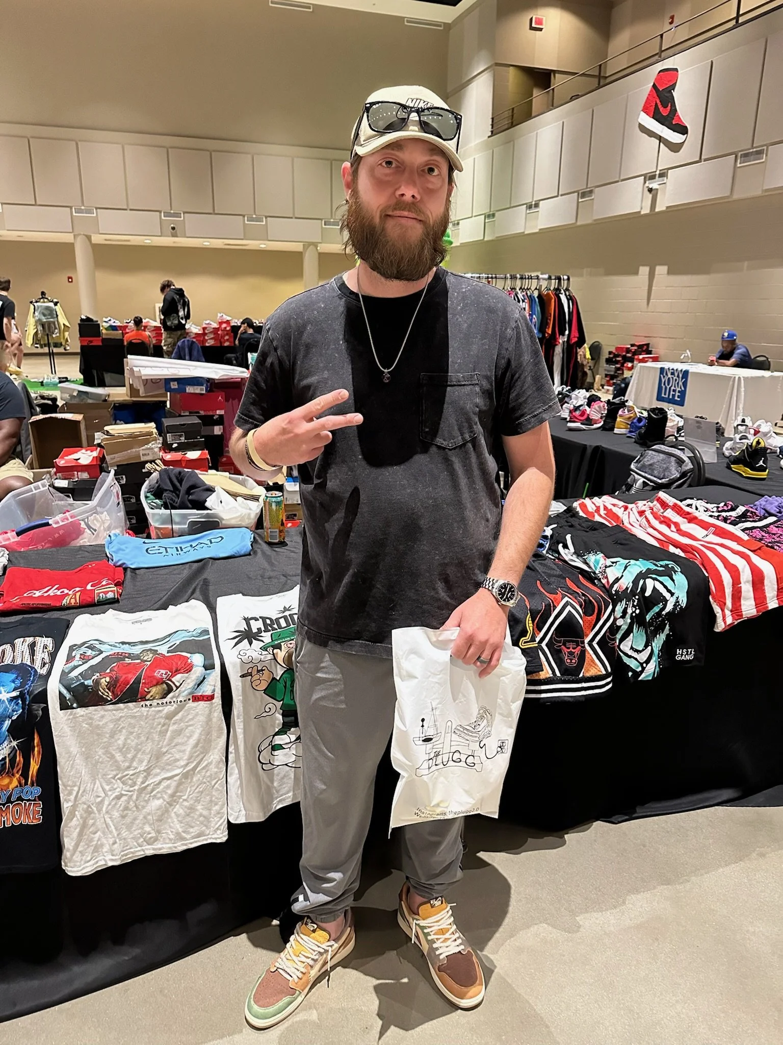 A man with a beard and casual clothing standing in an indoor market or swap meet, holding a white shopping bag, with clothing and sneakers on display behind him.