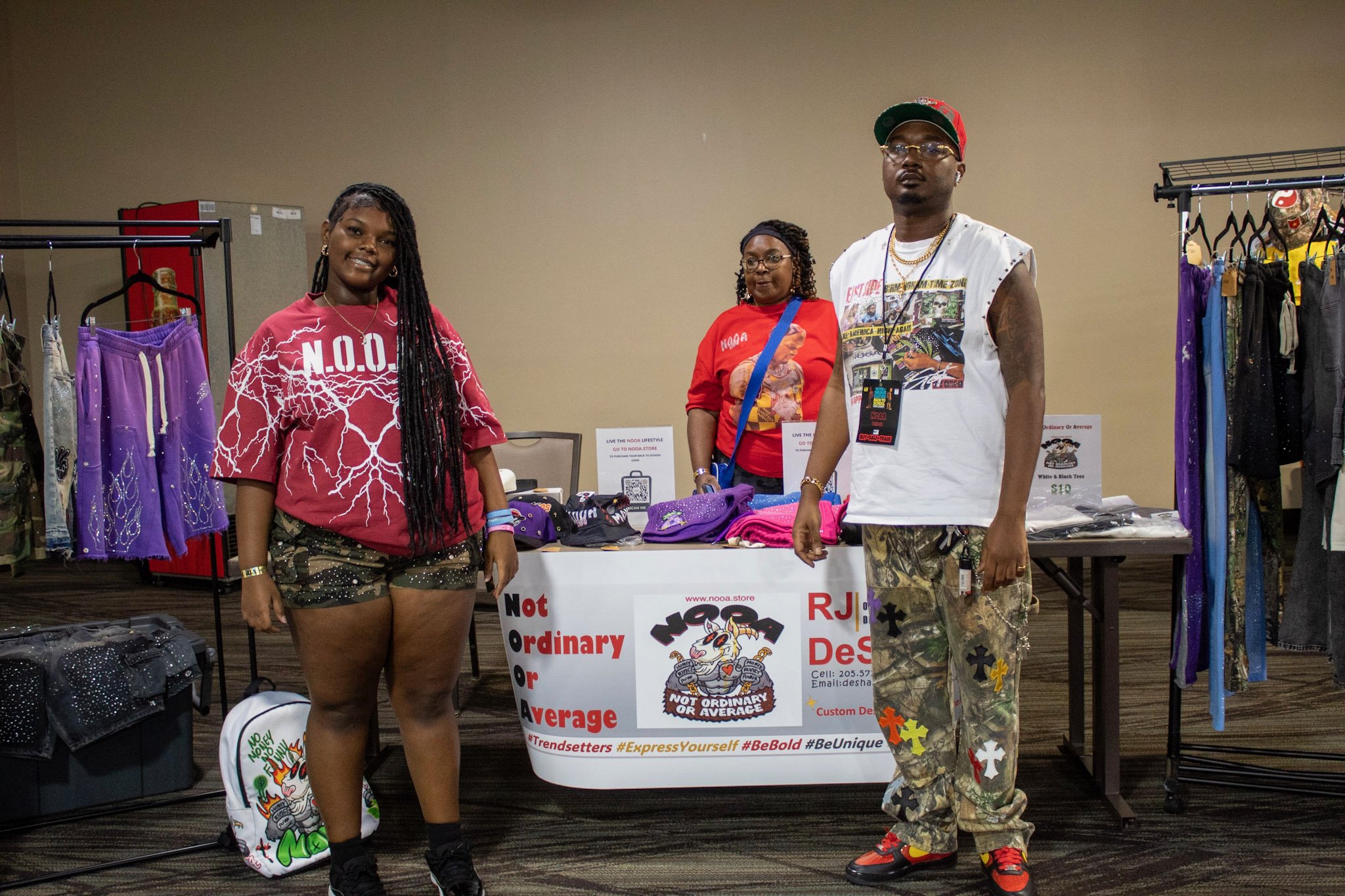Two women and a man at a merchandise booth, with clothing and accessories on racks and tables, in an indoor event.