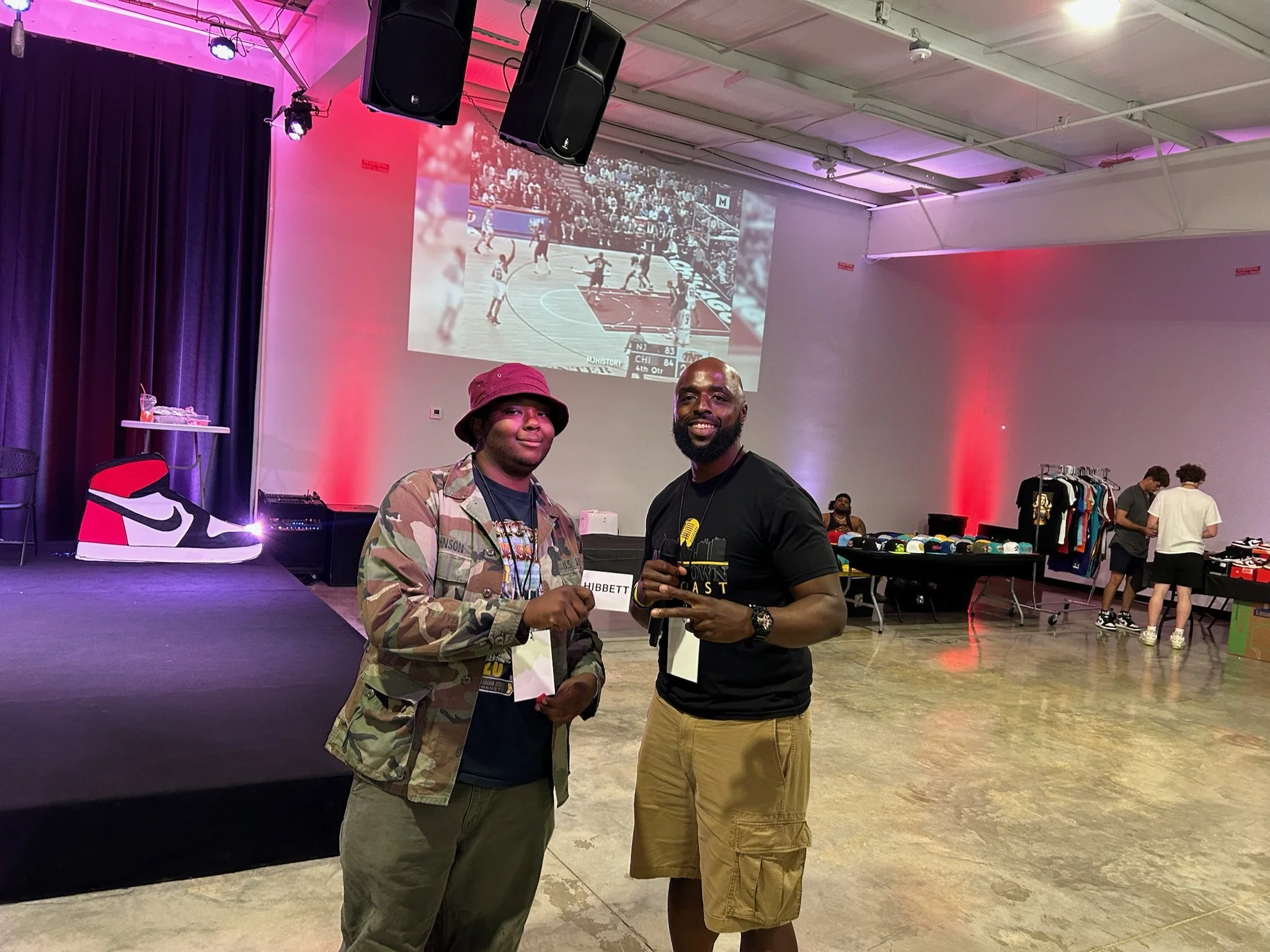 Two men stand together in a large room with a high ceiling, pink and red lighting, and a stage with a giant sneaker display on the left. One man wears a camouflage jacket and bucket hat, holding a piece of paper, while the other wears a black T-shirt