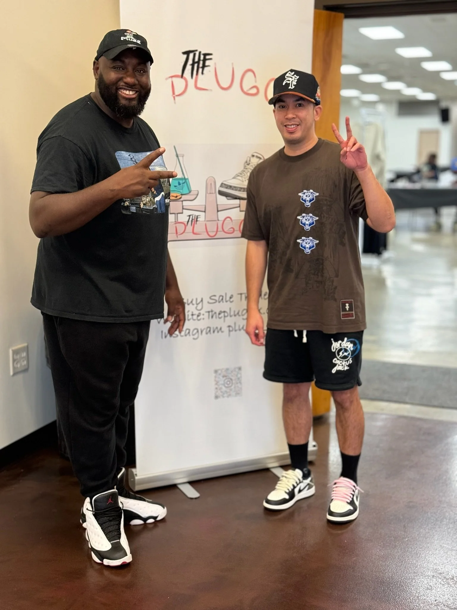 Two men standing inside a building in front of a sign for 'The Plug.' One man is wearing a black t-shirt and black pants, pointing with his right hand and smiling. The other man is wearing a brown t-shirt, black shorts, and multicolored sneakers, mak