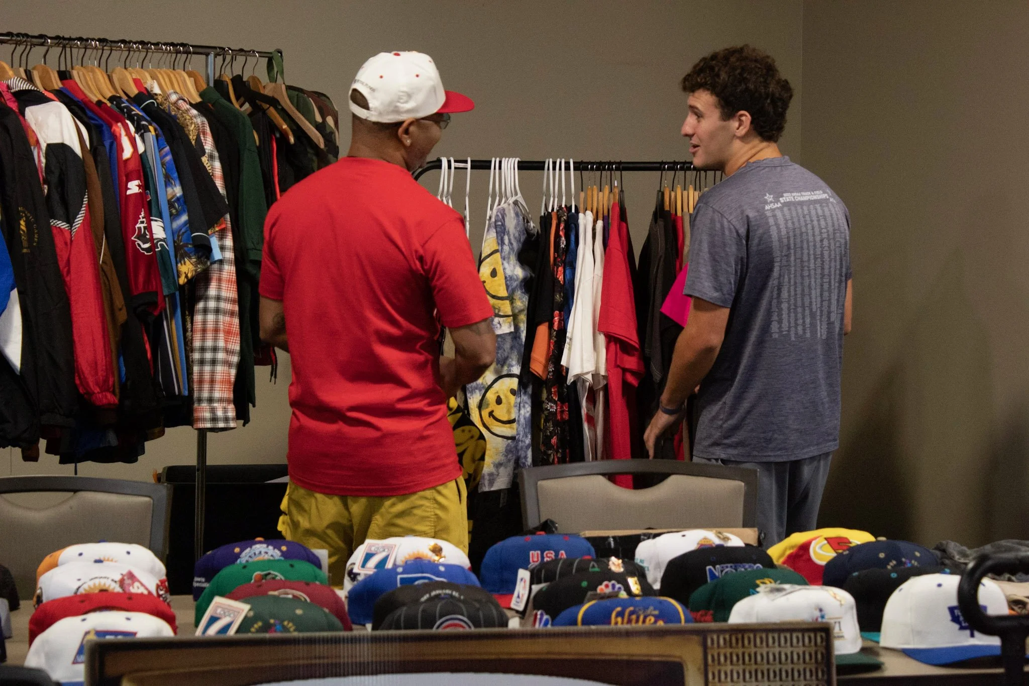 Two men shopping for clothes and hats at a store. One man is wearing a red t-shirt and yellow shorts with a white and red cap, talking to the other man in a gray t-shirt. Clothing racks with various shirts and jackets are behind them, and tabletop di