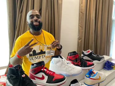 Man with a beard and wearing a yellow T-shirt displaying sneakers, sneakers placed on a table, in a room with beige curtains.