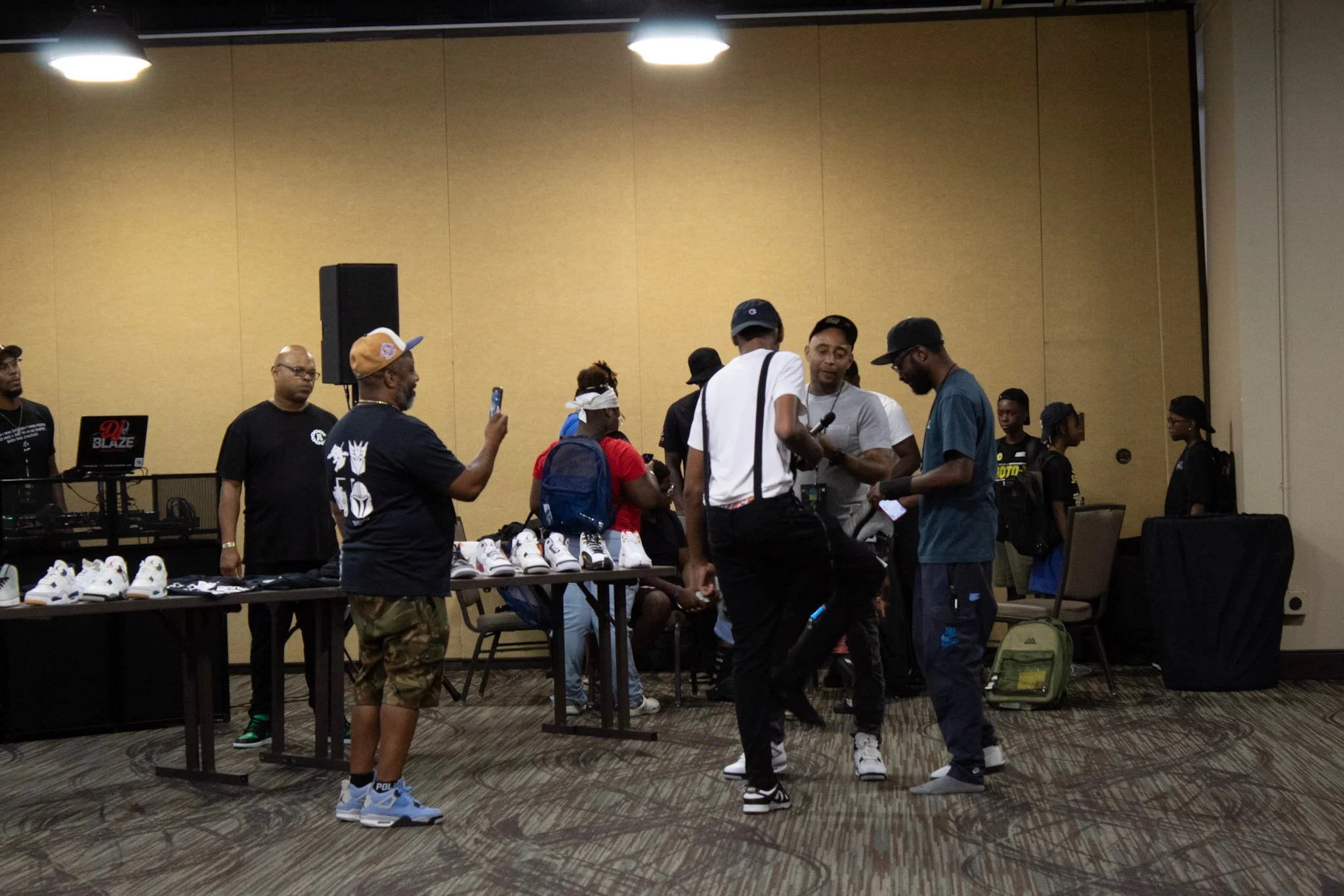 Group of people gathered around tables with white sneakers displayed, in an indoor event space with beige walls and overhead lights.