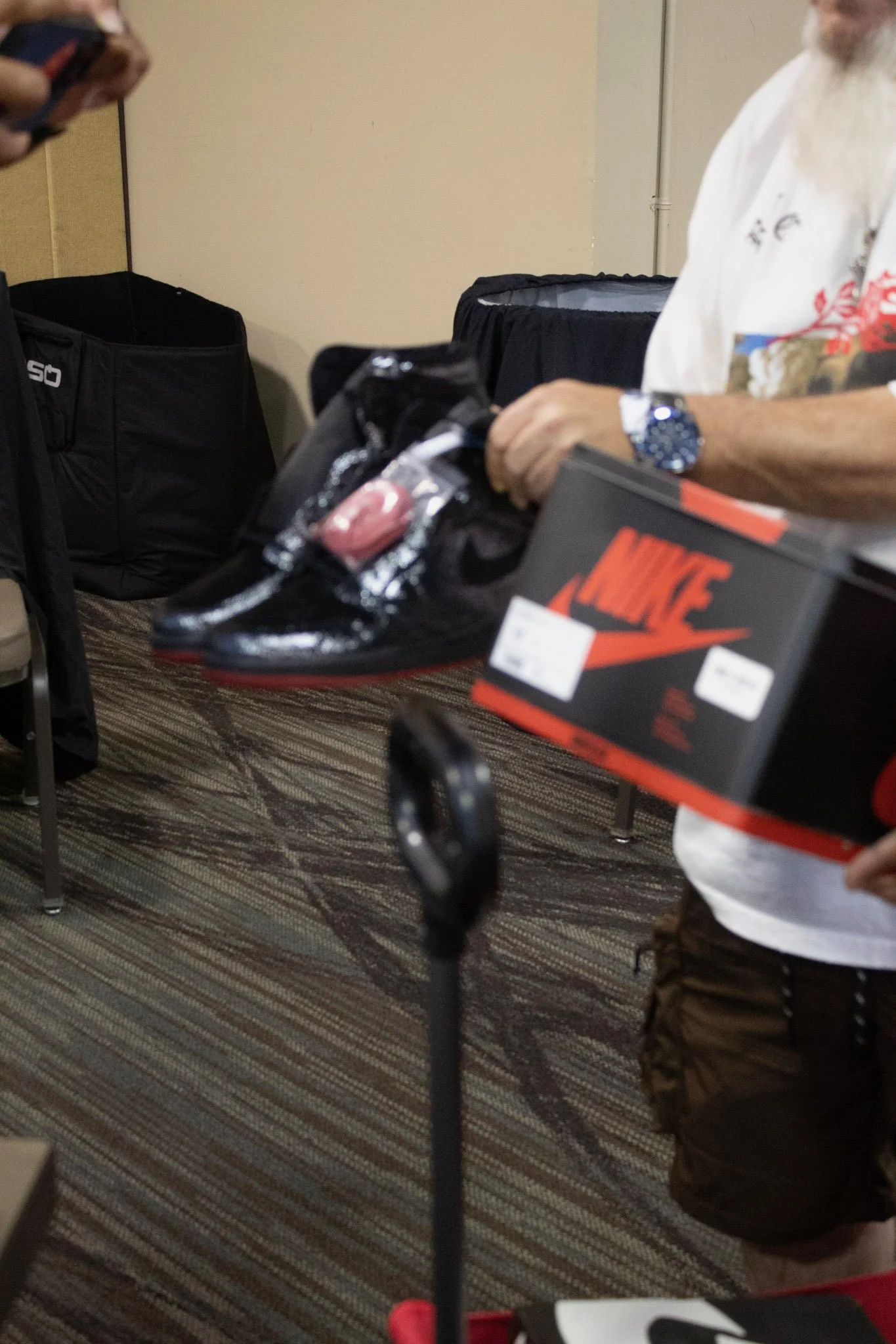 Person holding a pair of black and red Nike sneakers with a pink tag attached and a Nike shoebox in front of them, in an indoor setting.