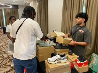Two men exchanging items at a table with shoes and boxes in a hotel conference room.