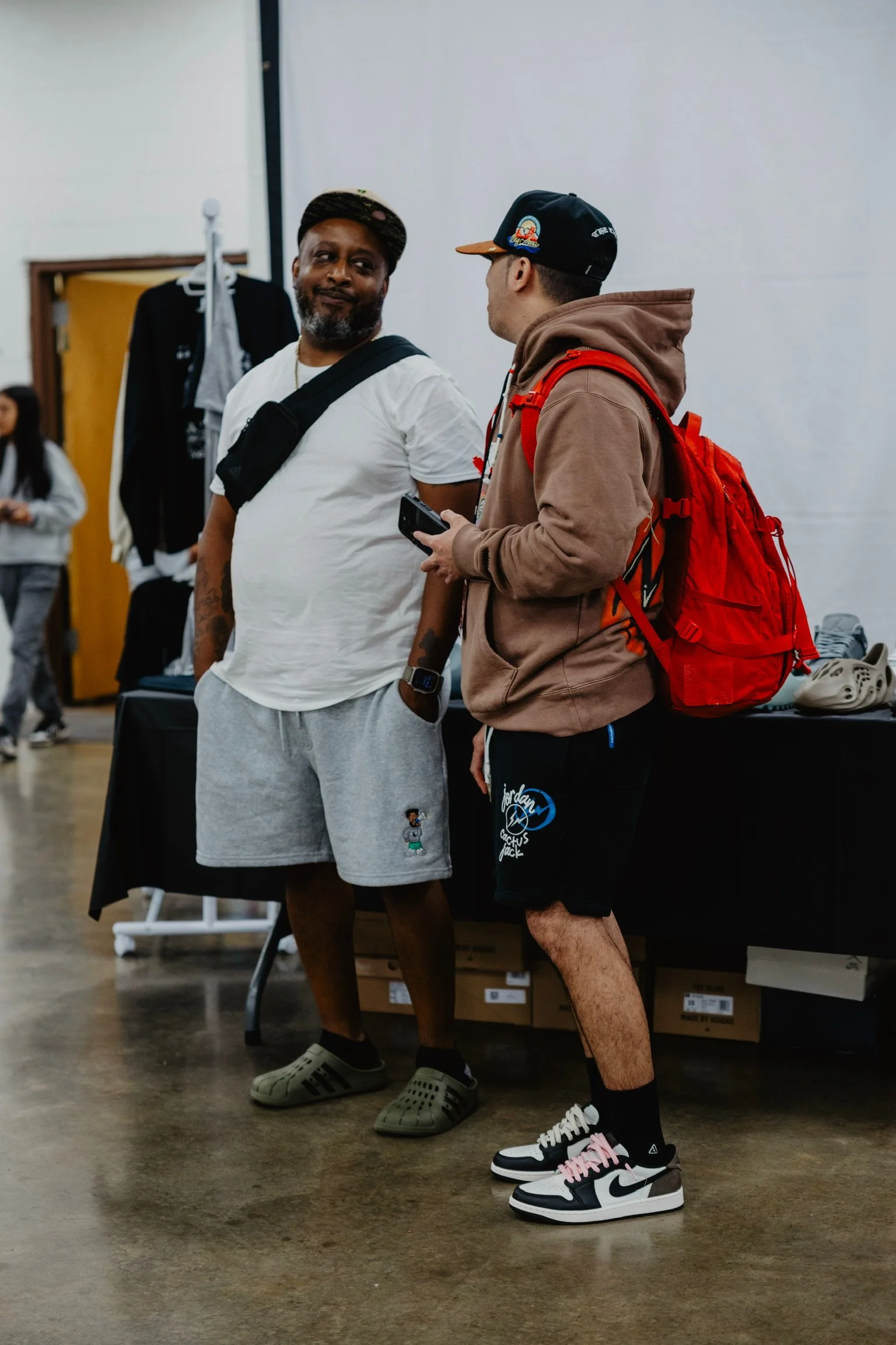 Two men talking inside a room with clothing displays in the background. One man is wearing a white t-shirt, gray shorts, and green Crocs. The other man is wearing a brown hoodie, black shorts, and Nike sneakers, with a red backpack.