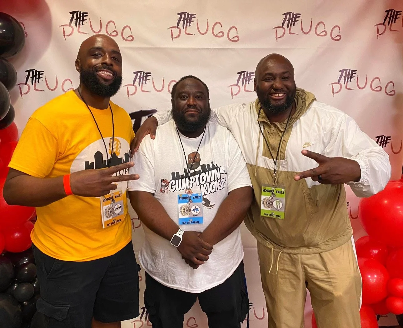 Three men standing together at an event, smiling at the camera. They are wearing event badges around their necks. The background features a wall with the repeated logo 'The Deugg' and balloon decorations in black and red.