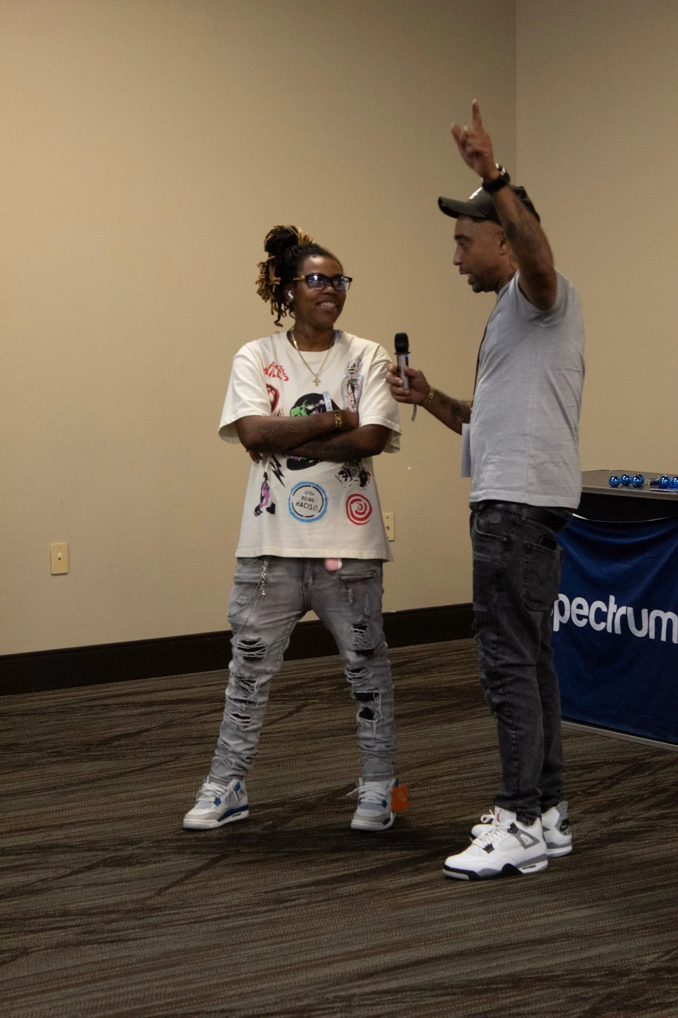Two people conversing in a room with a plain wall. One person is holding a microphone, wearing a gray T-shirt and black jeans, while the other is wearing a graphic T-shirt and distressed jeans.