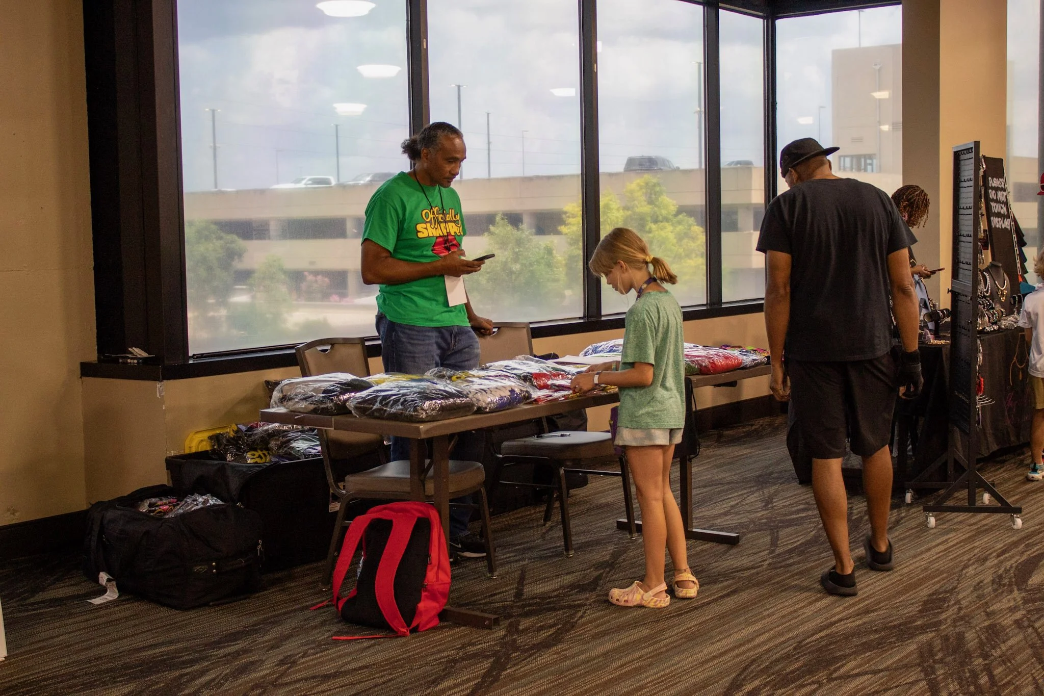 A man in a green T-shirt and a girl in a green T-shirt are looking at merchandise on a table next to a man in a black t-shirt and a woman in a light-colored shirt at an indoor event.