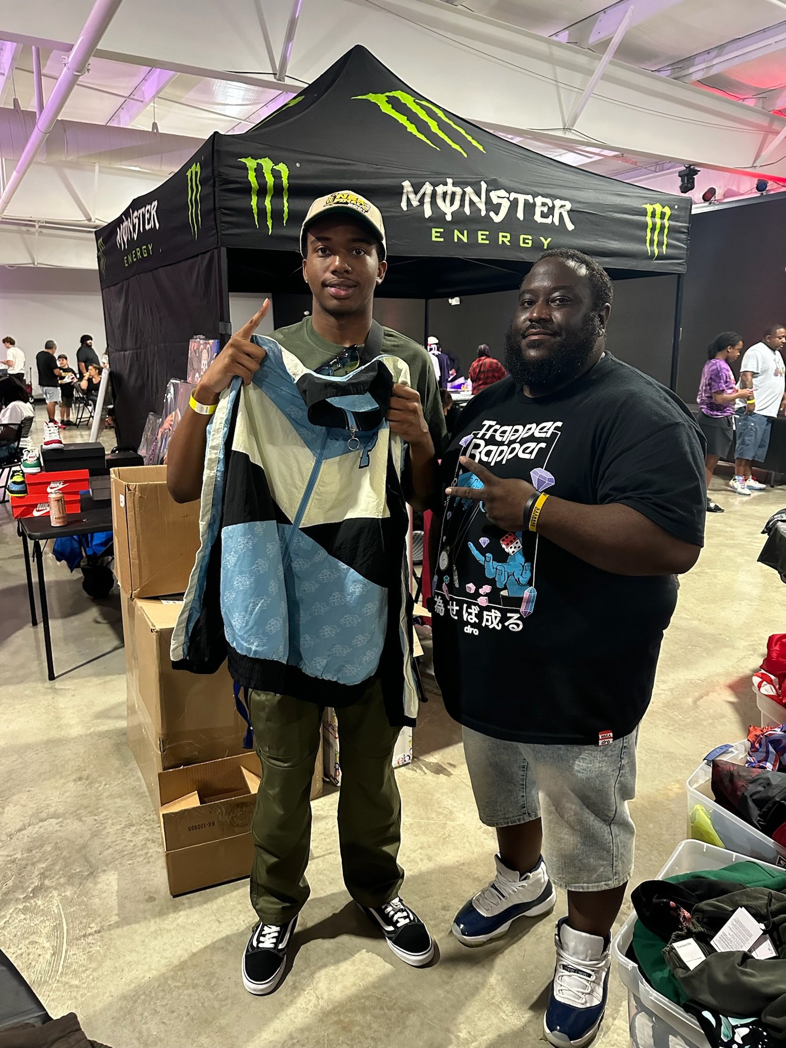 Two men standing in front of a black Monster Energy tent at an indoor event, one holding a jacket and the other making a peace sign.
