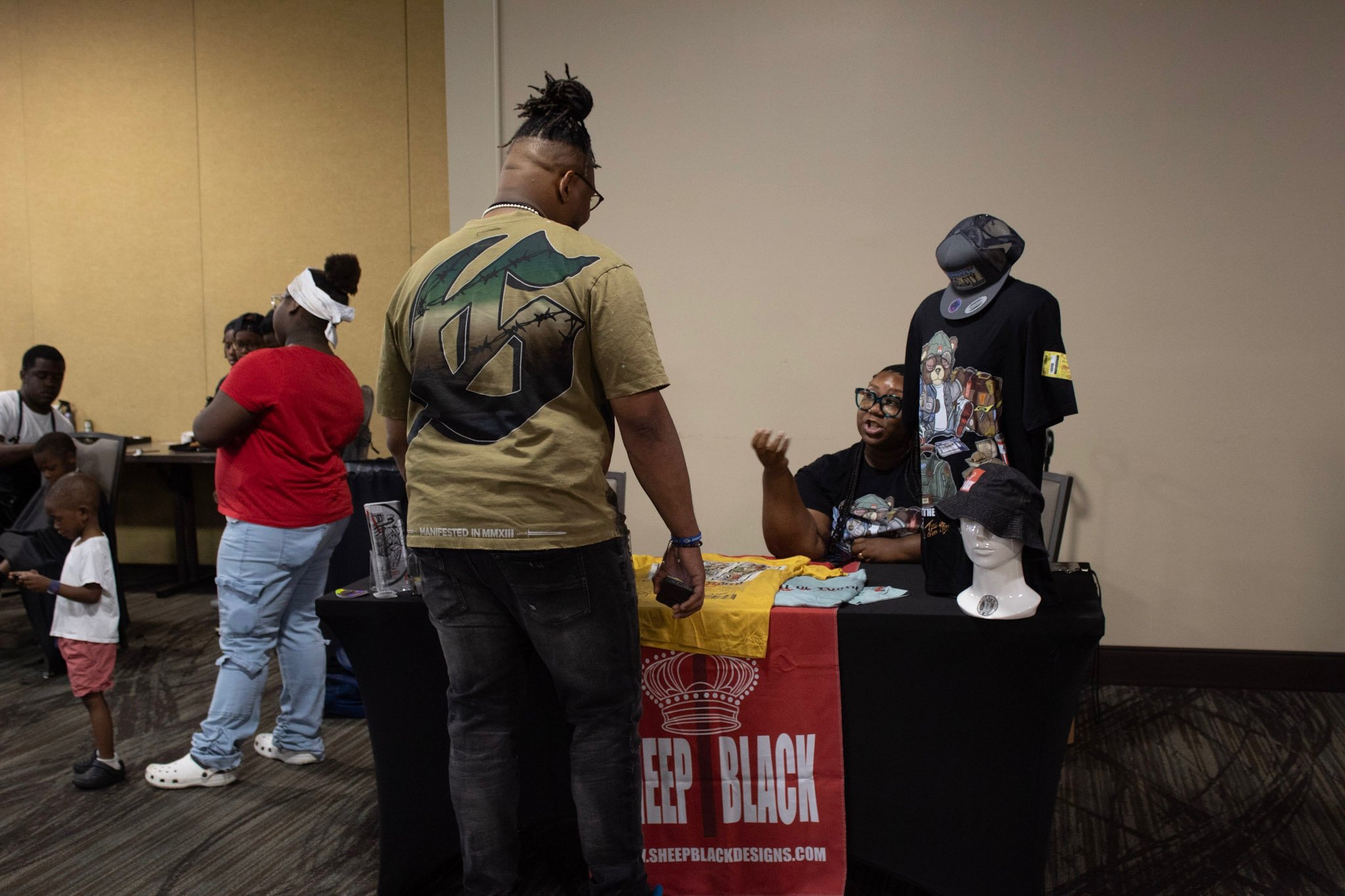 A booth at an event displaying t-shirts and hats, with a woman seated behind the table talking to a man standing in front. The table has a red banner that reads 'SHEEP BLACK' and features various merchandise, including headwear and small accessories.