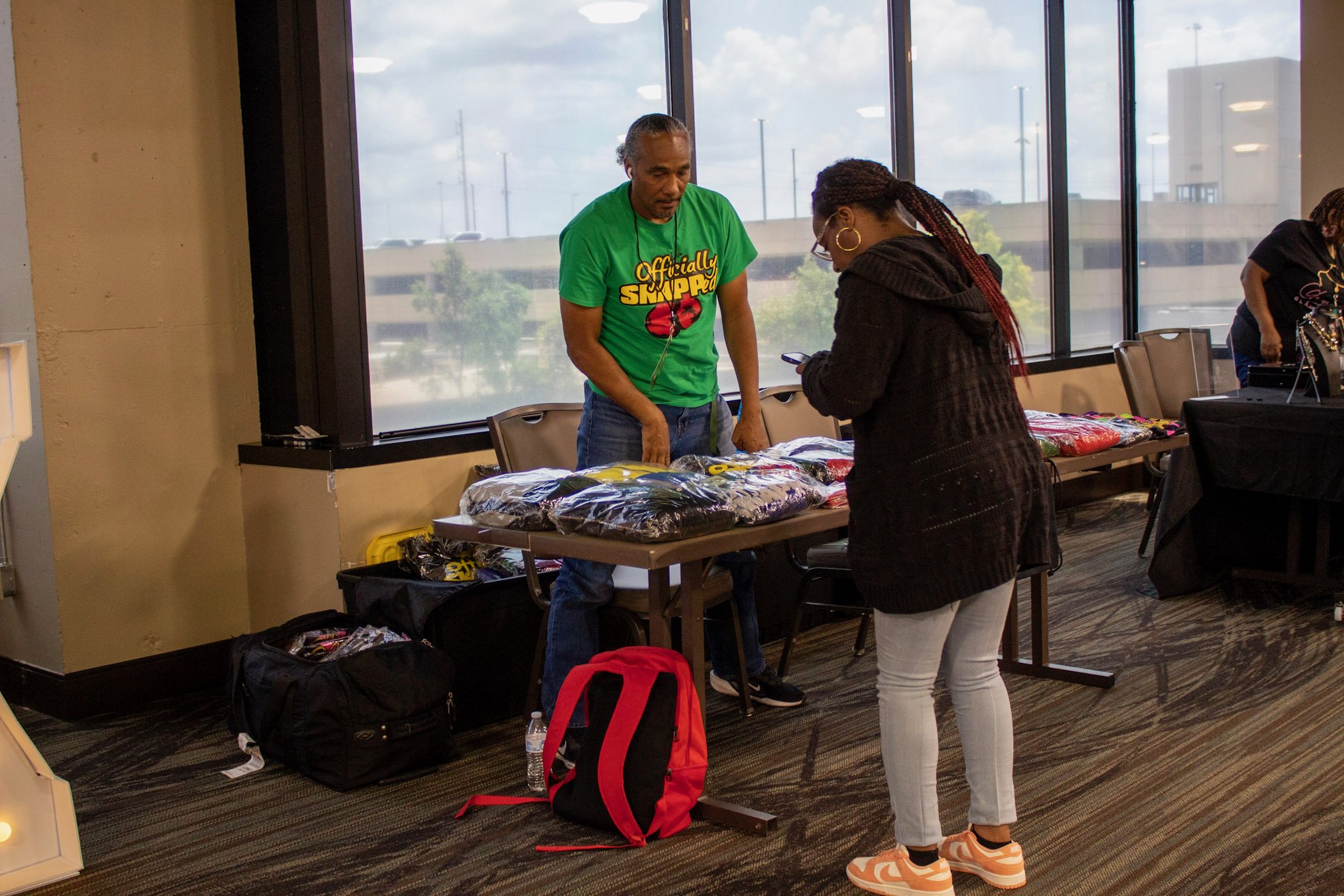 A man and a woman at a table with folded clothing items inside a room with large windows. The man is wearing a green t-shirt, and the woman is wearing a black hoodie and white pants.