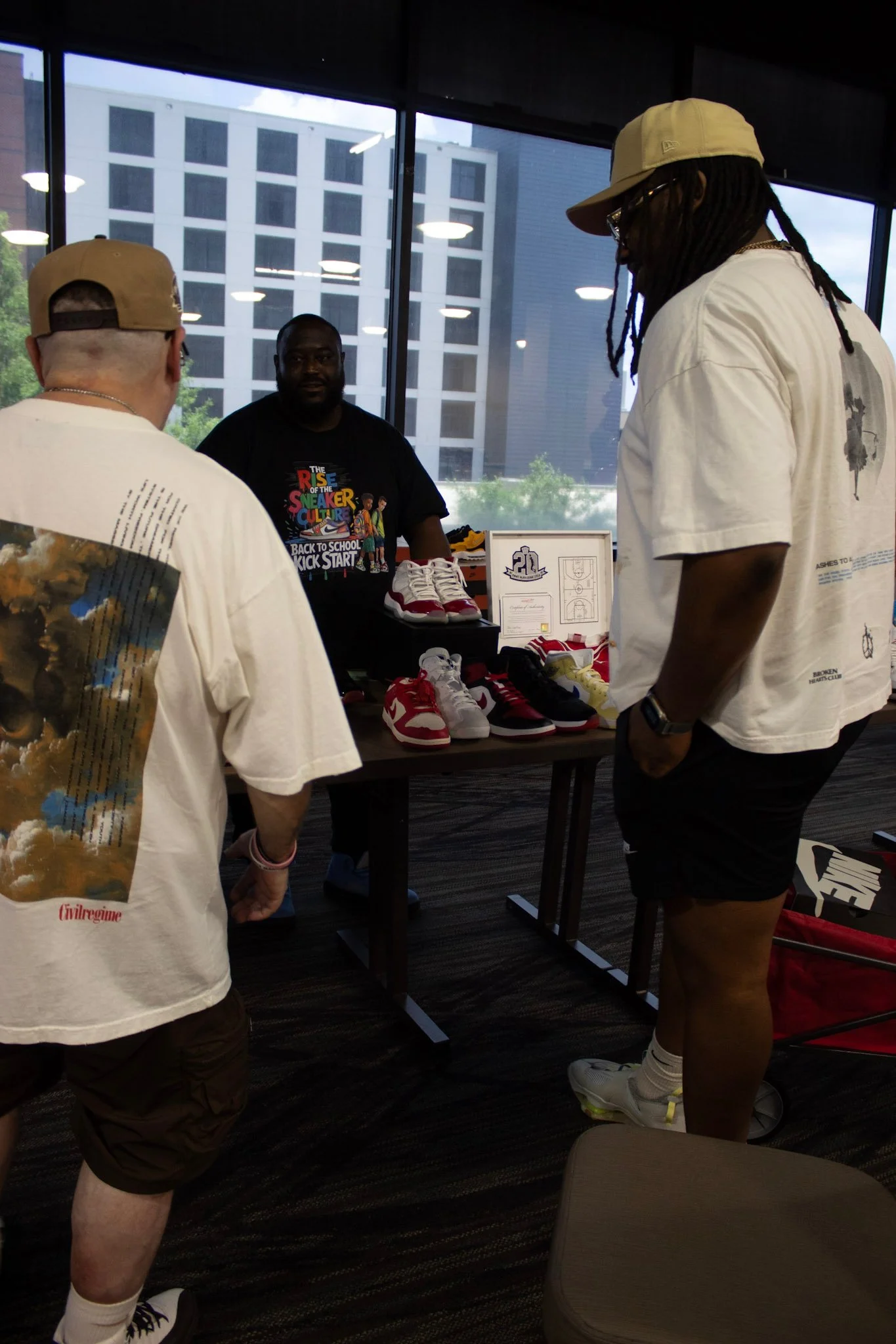 Three men are at a sneaker booth indoors, with large windows behind them showing a cityscape. The booth has various sneakers on display, including red, white, black, and yellow shoes. The man behind the booth is wearing a black T-shirt with colorful 