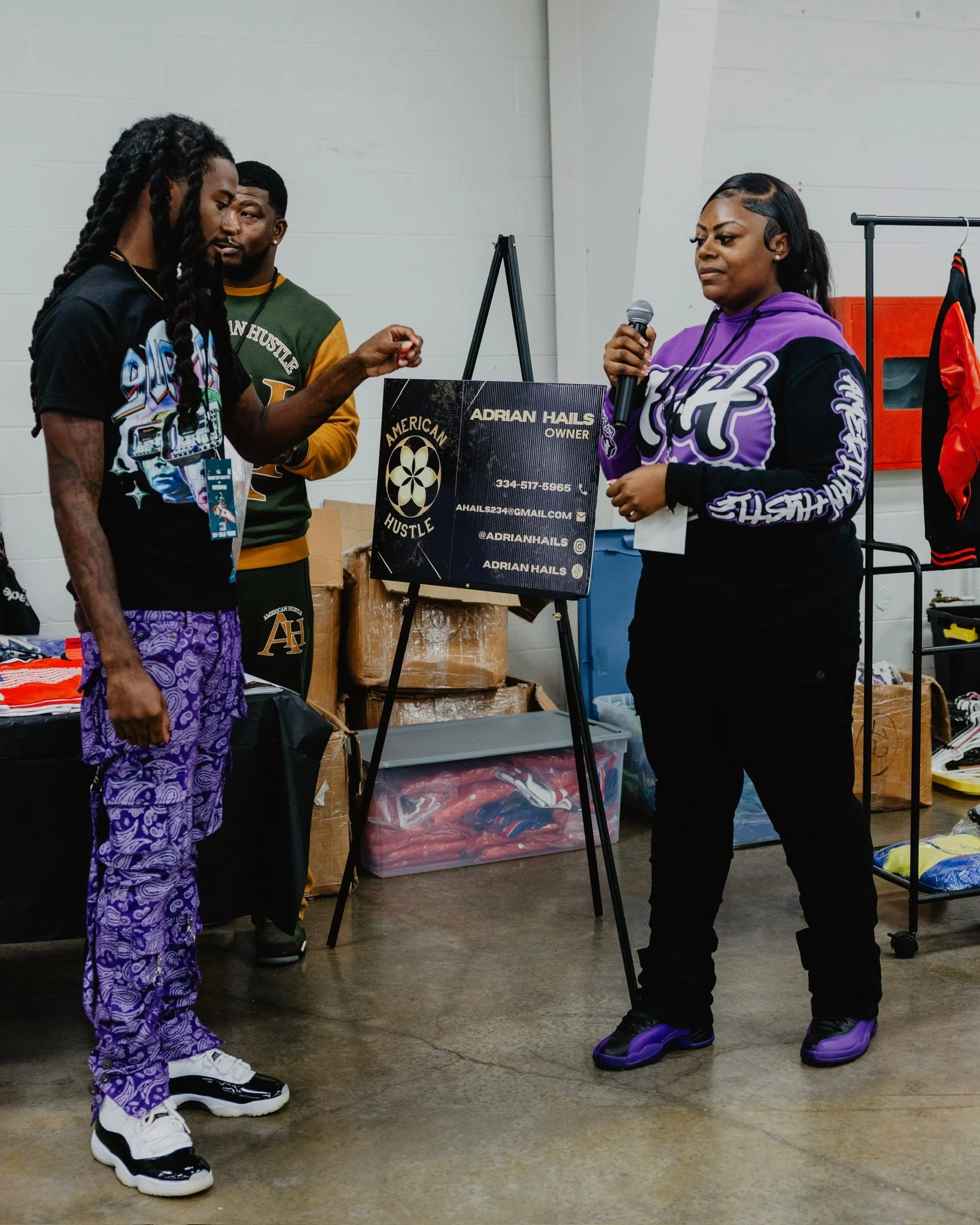 Three people at a booth with a sign that reads 'Adrian Hails Owner,' at an indoor event. One person with long dreadlocks, wearing a graphic t-shirt and purple pants, is talking with a woman holding a microphone, who is wearing a purple and black hood